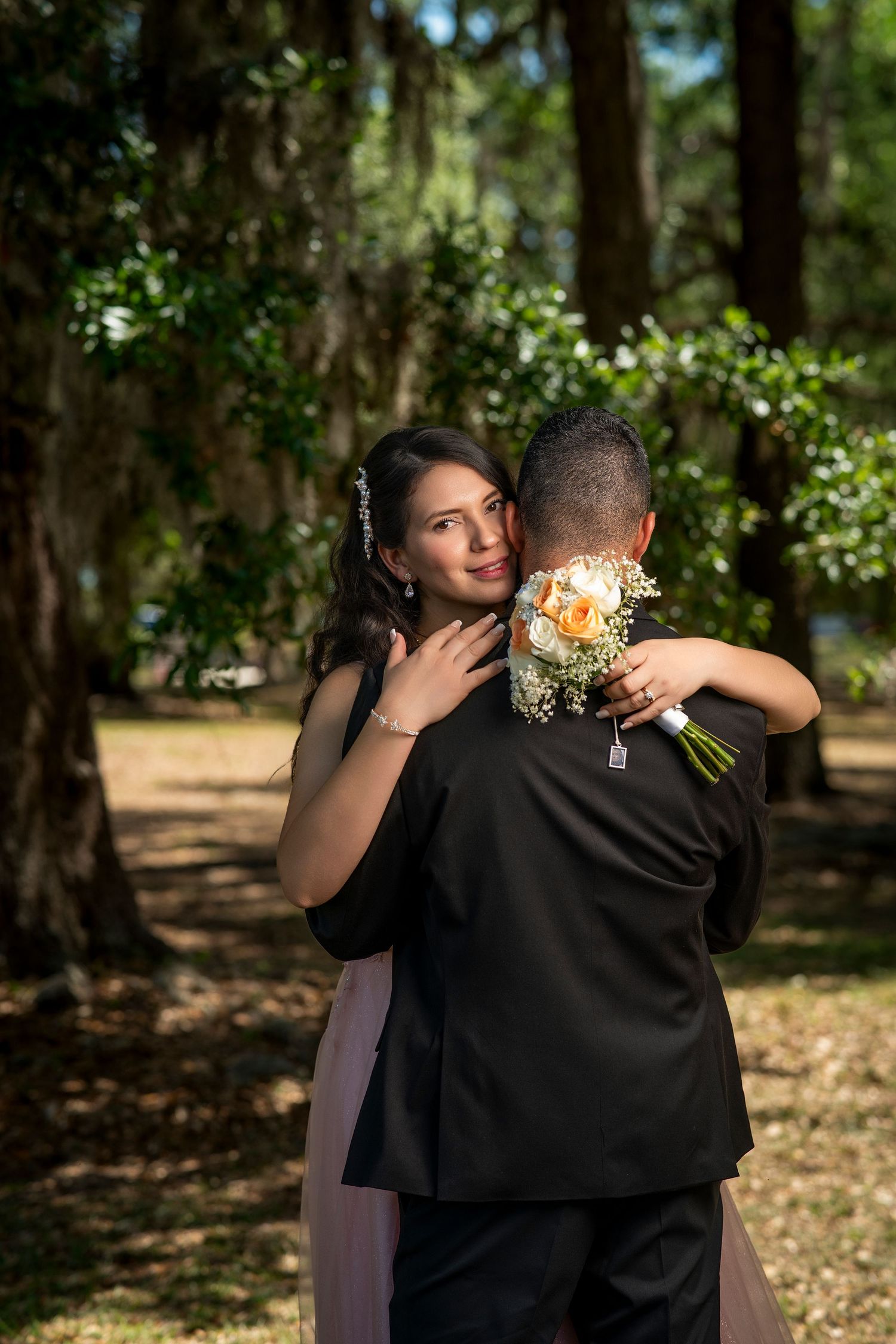 Bride holding bouquet embraces groom in black suit outdoors among lush green trees in romantic wedding portrait.