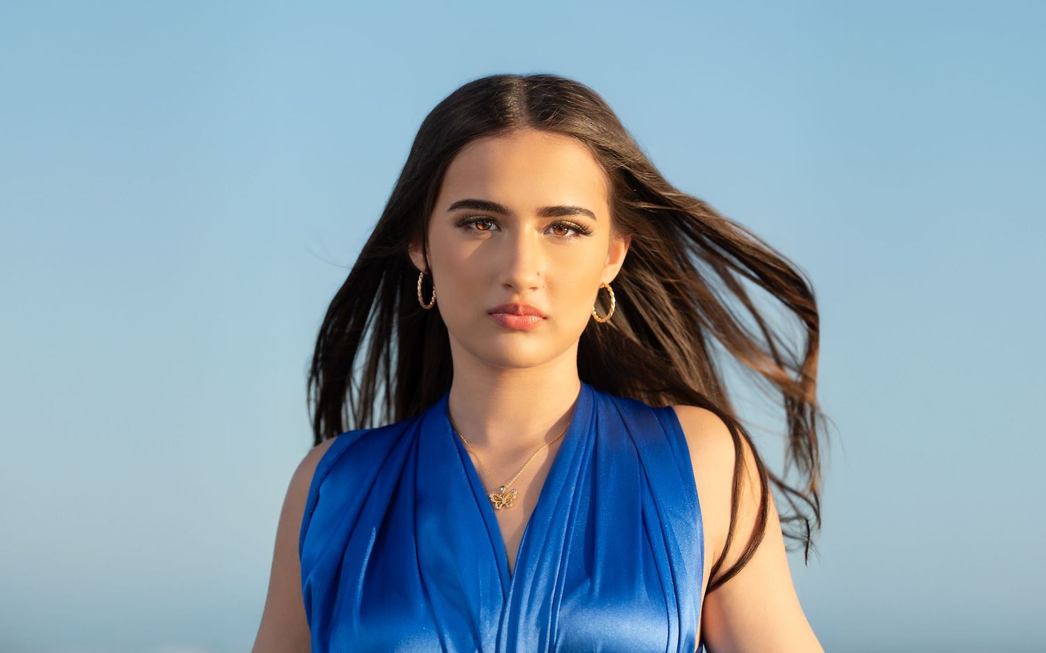 Woman in bright blue sleeveless top poses against soft blue sky background with wind-swept dark hair.