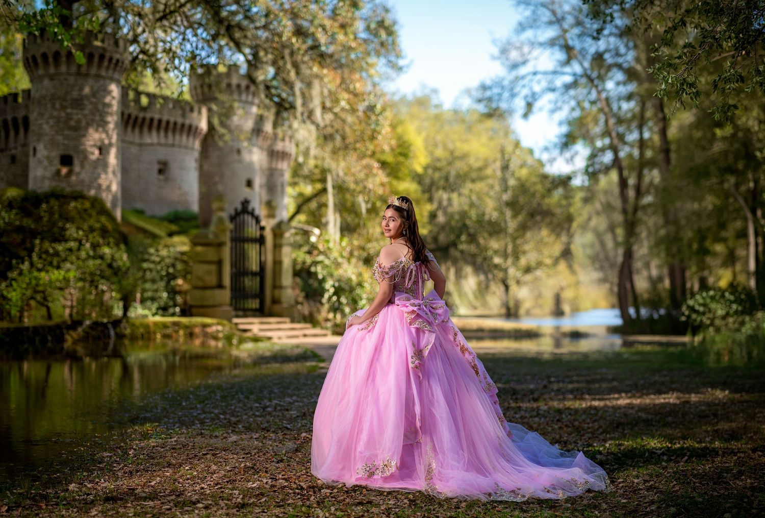 Young woman in pink ball gown poses before a castle surrounded by lush greenery and a serene pond.