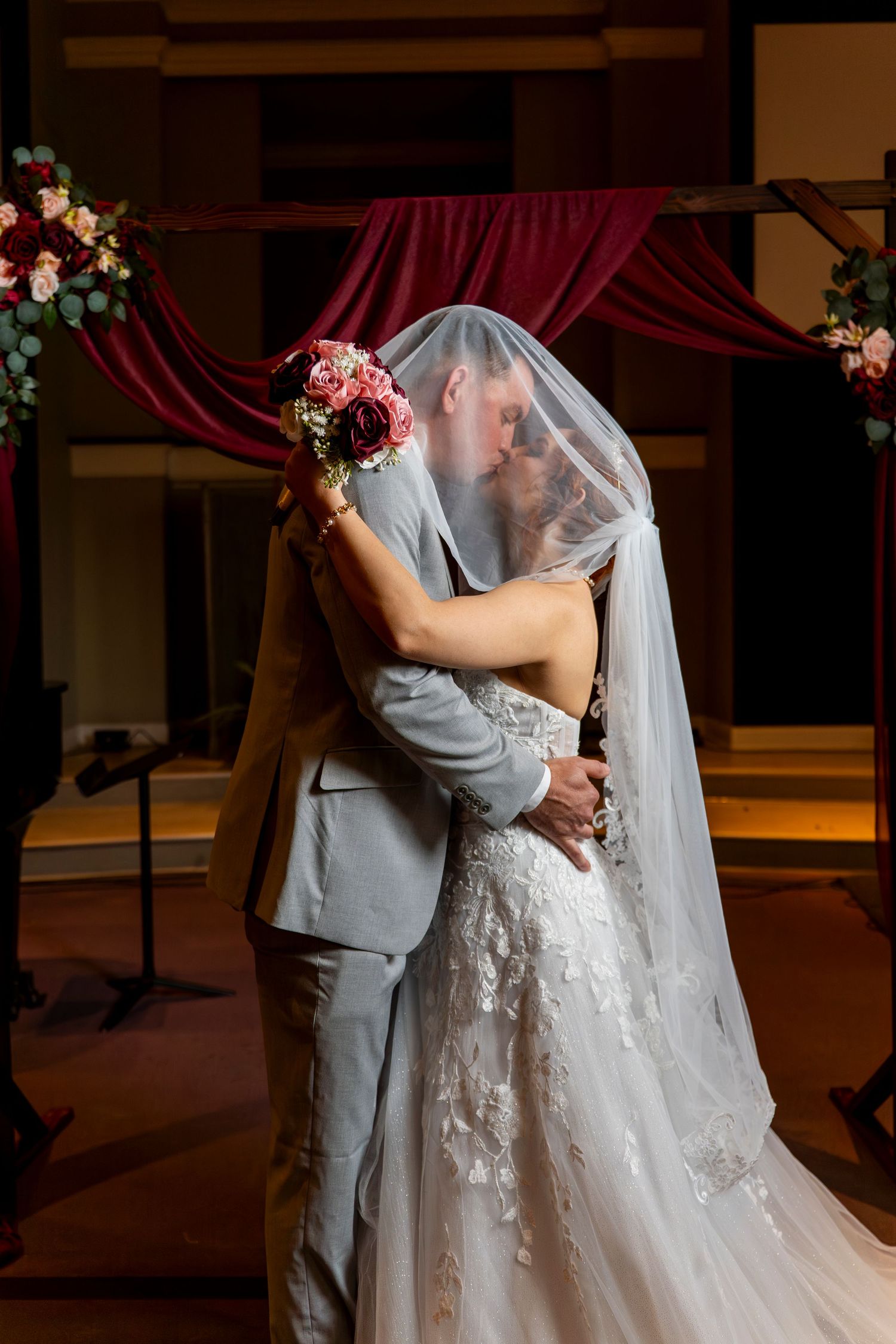 A romantic wedding couple embraces on a dance floor with elegant red drapes and floral decorations in the background.