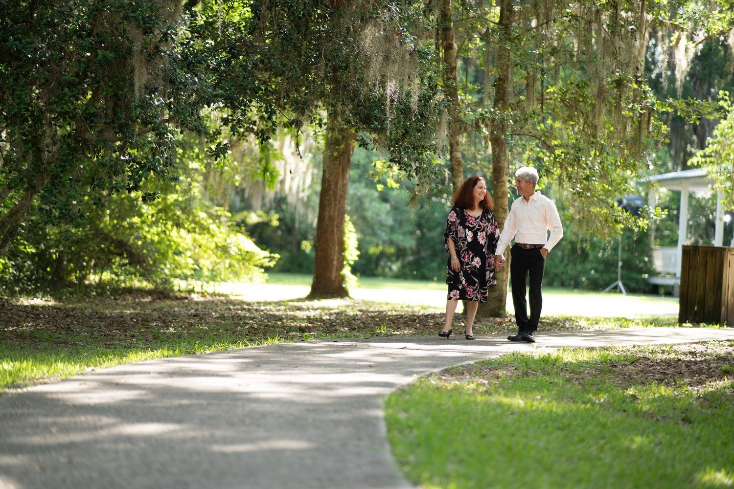 A couple takes a leisurely stroll along a sunlit path beneath Spanish moss-draped trees in a serene park setting.