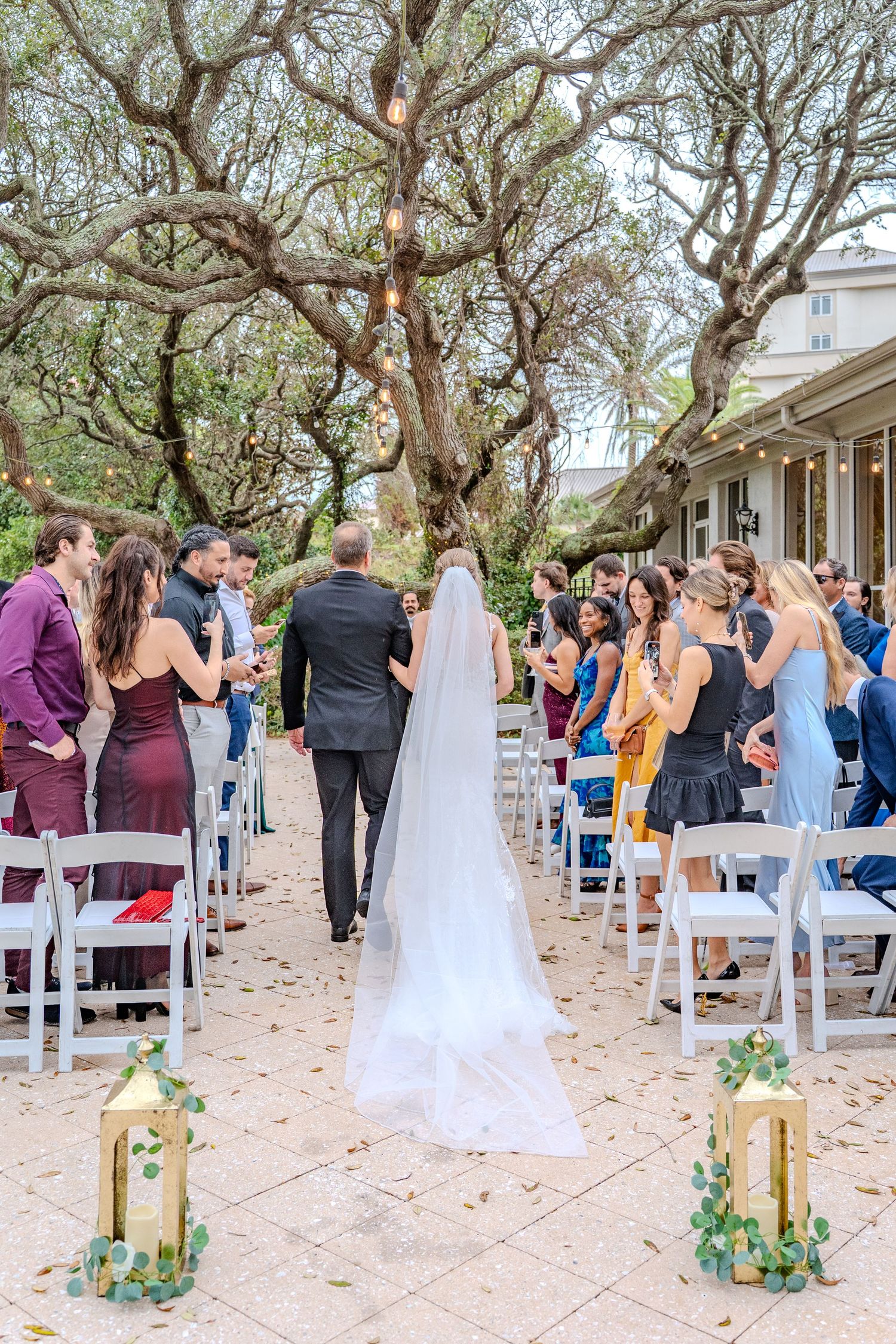 Outdoor wedding ceremony setup with white chairs under canopied trees decorated with floral arrangements.