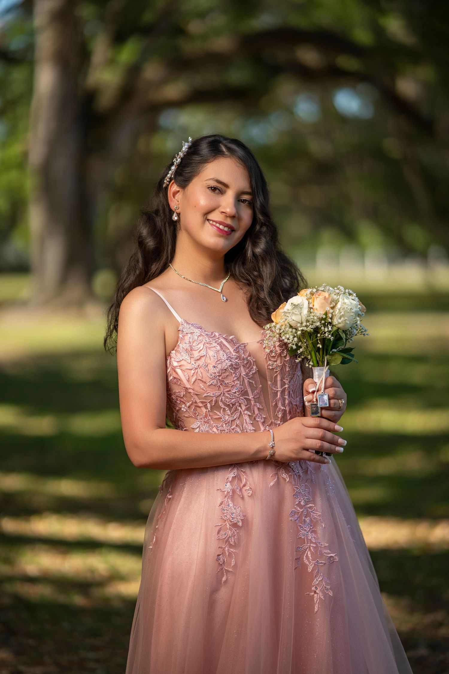 A smiling young woman in a blush pink lace gown holds a white floral bouquet in a sunlit outdoor setting.