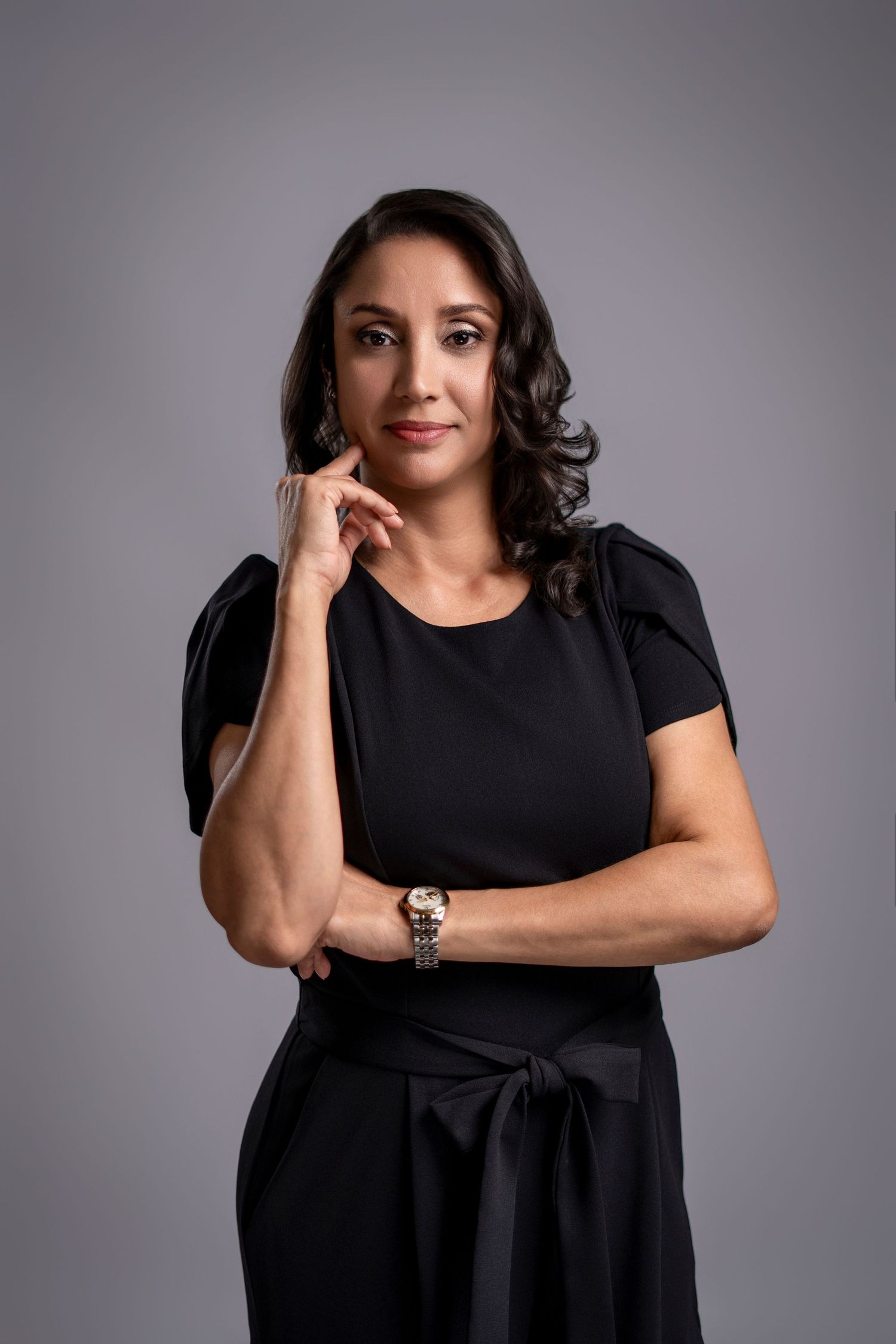 Professional woman in black dress posing confidently with hand on chin against gray background.