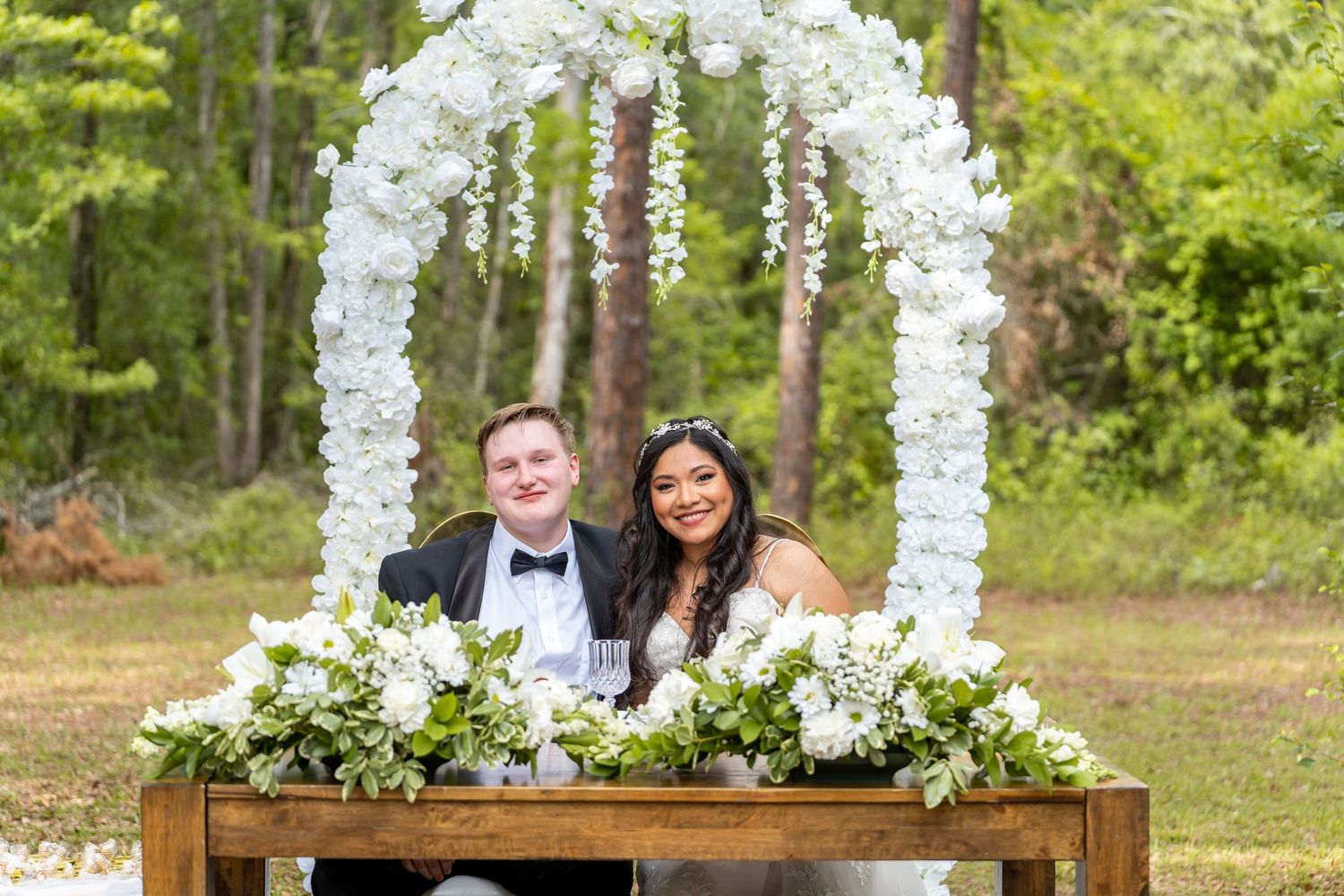 Newlywed couple smiling at sweetheart table under white floral arch with lush greenery backdrop at outdoor wedding.