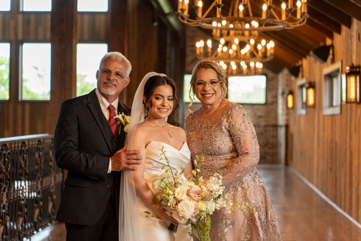 Bride posing with parents in elegant wedding venue hallway with chandelier lighting.