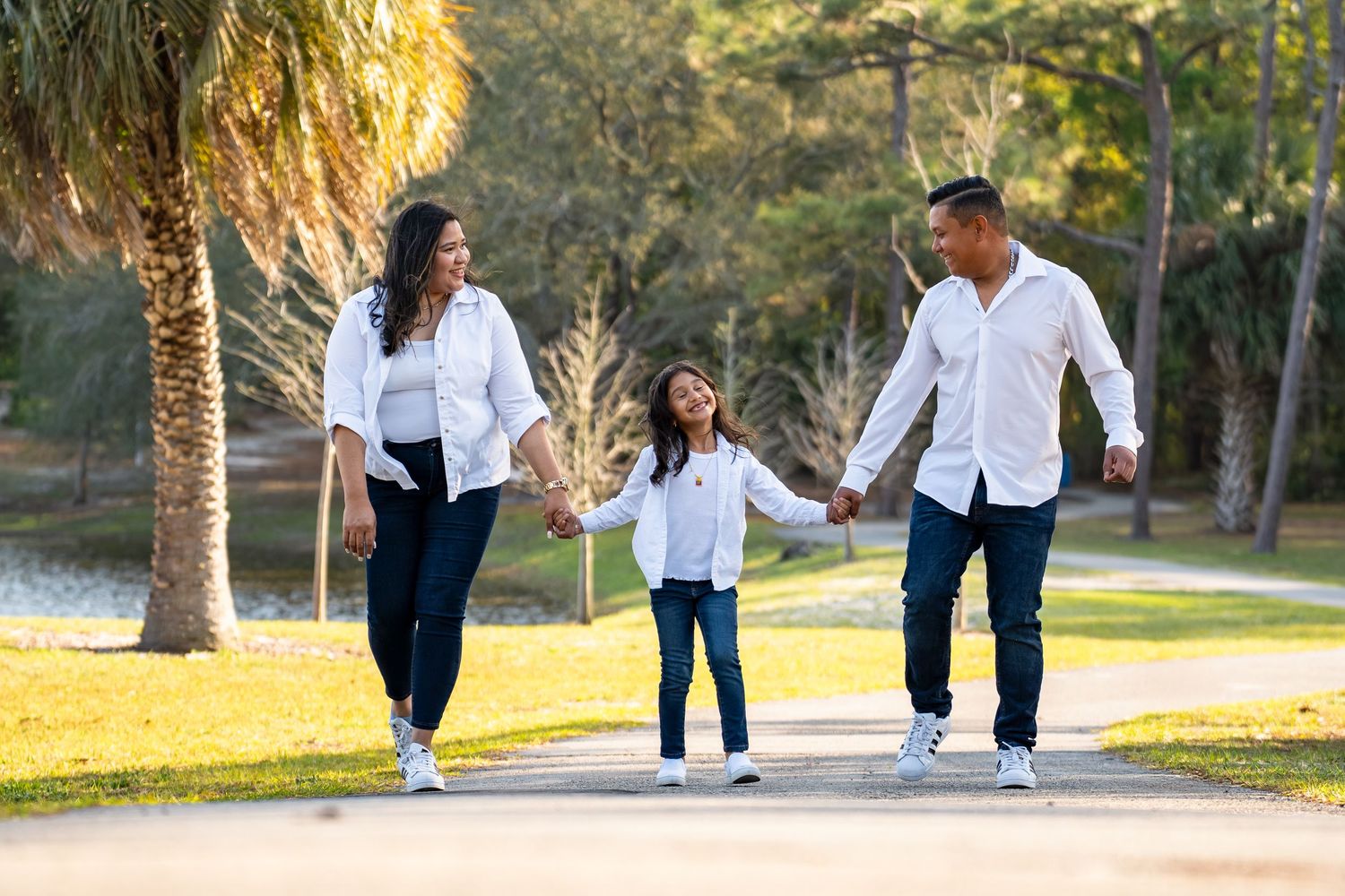 Family wearing casual white outfits walking together on tree-lined path during golden sunset hour.
