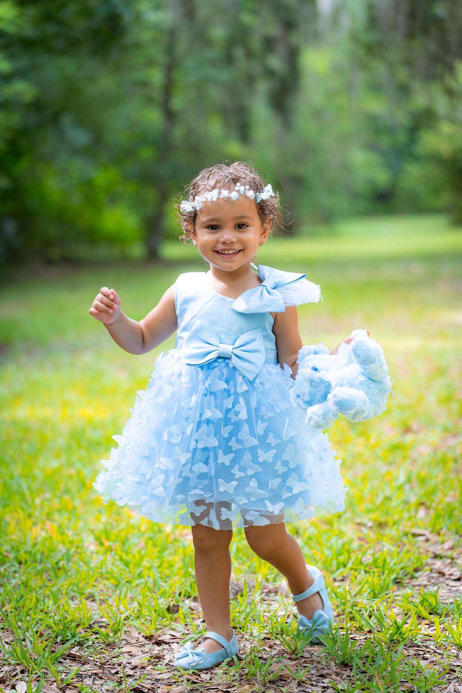 A child wearing a light blue ruffled dress and floral crown stands in a grassy outdoor setting.
