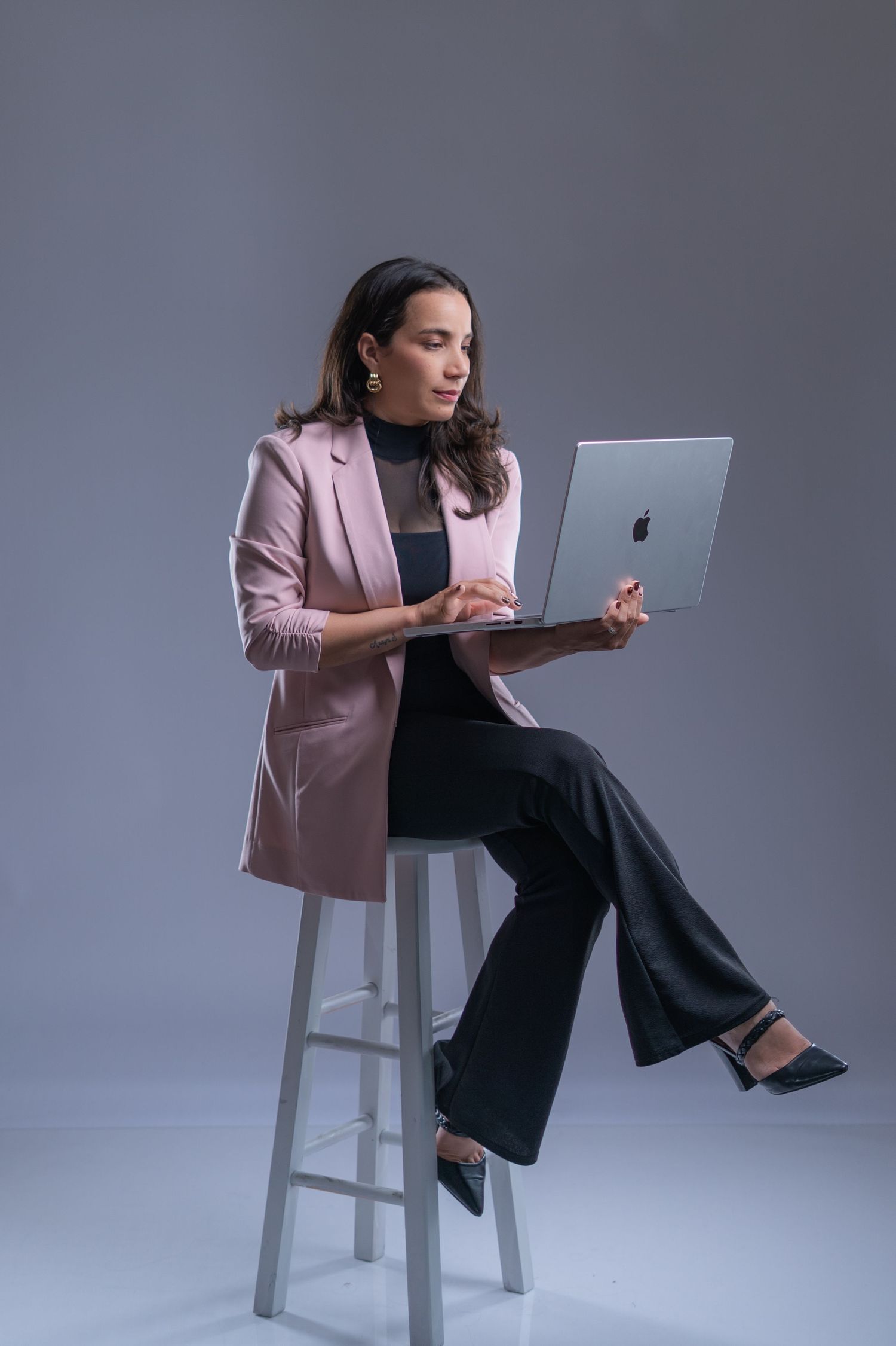 Professional wearing pink blazer and black pants works on laptop while sitting on white stool in studio setting.