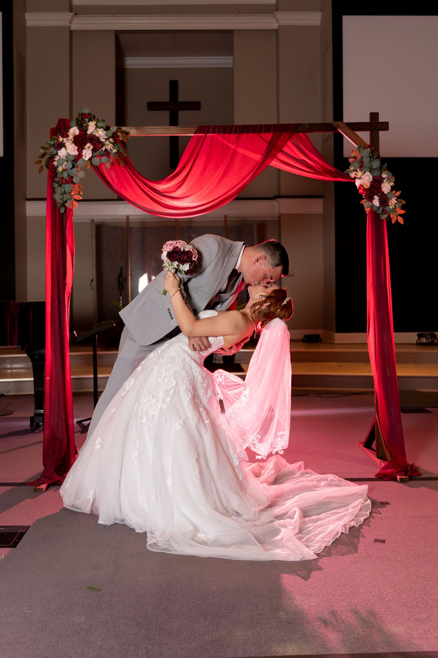 Wedding couple shares romantic dance under red drapery with dramatic lighting on pink carpeted floor.