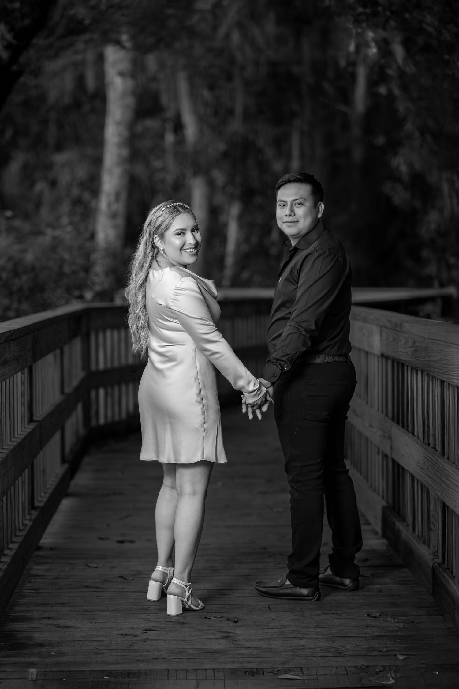 A couple poses together on a wooden bridge in black and white, holding hands and looking back at the camera.