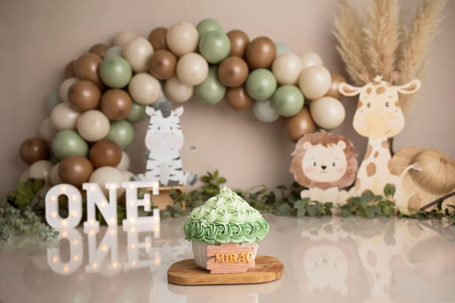 A neutral-toned first birthday party setup with balloon garland, pampas grass, and safari animal decorations in earthy colors.