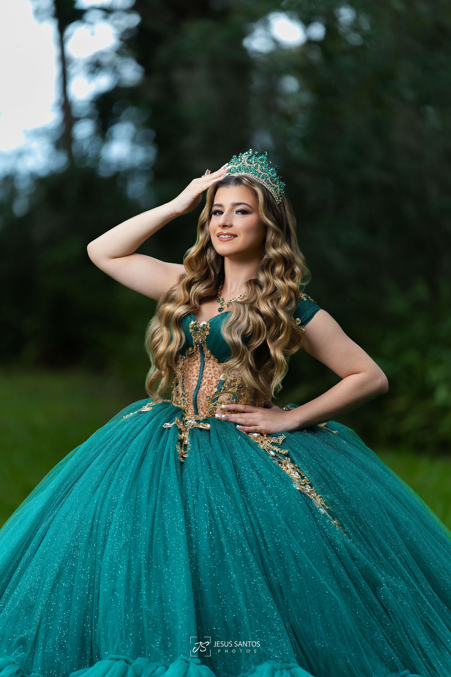 Young woman in sparkling emerald green quinceañera ball gown with gold accents and tiara poses outdoors.