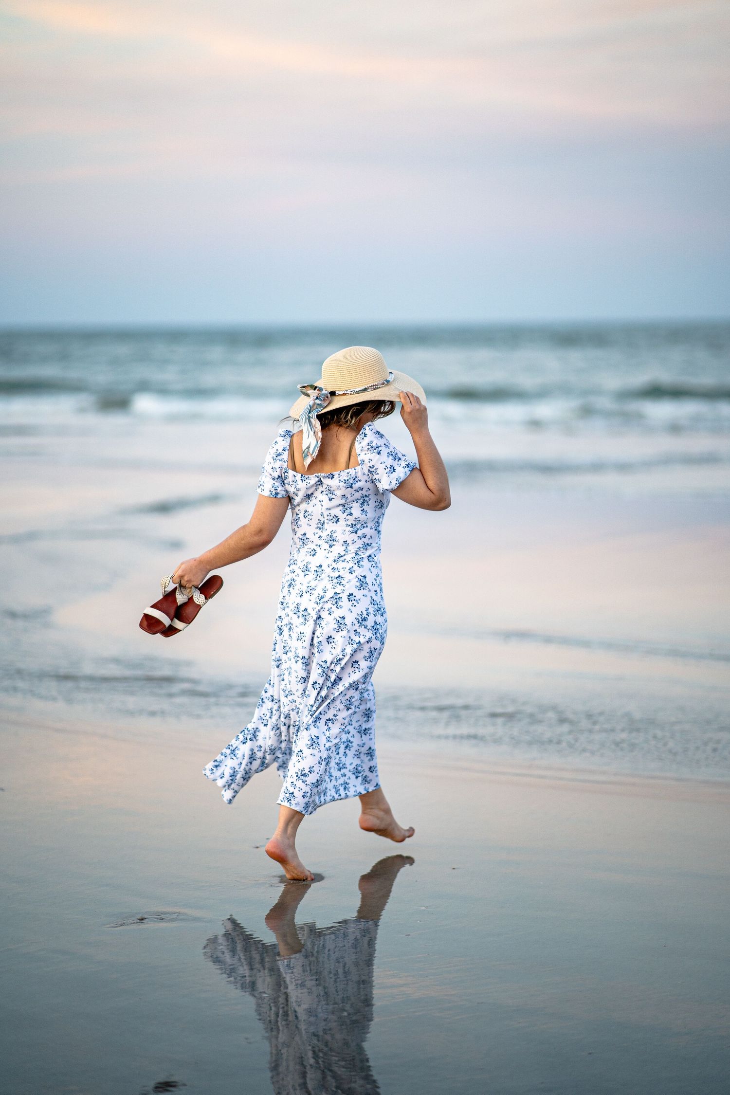Barefoot woman in flowy blue floral dress and sun hat walks along reflective beach at sunset holding sandals.