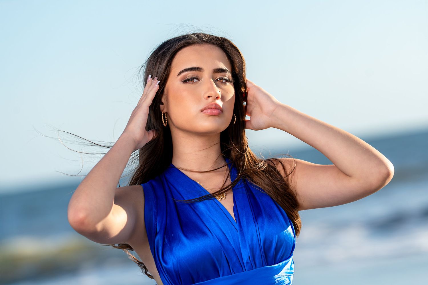 Woman in bright blue dress poses confidently on beach during sunset photoshoot.
