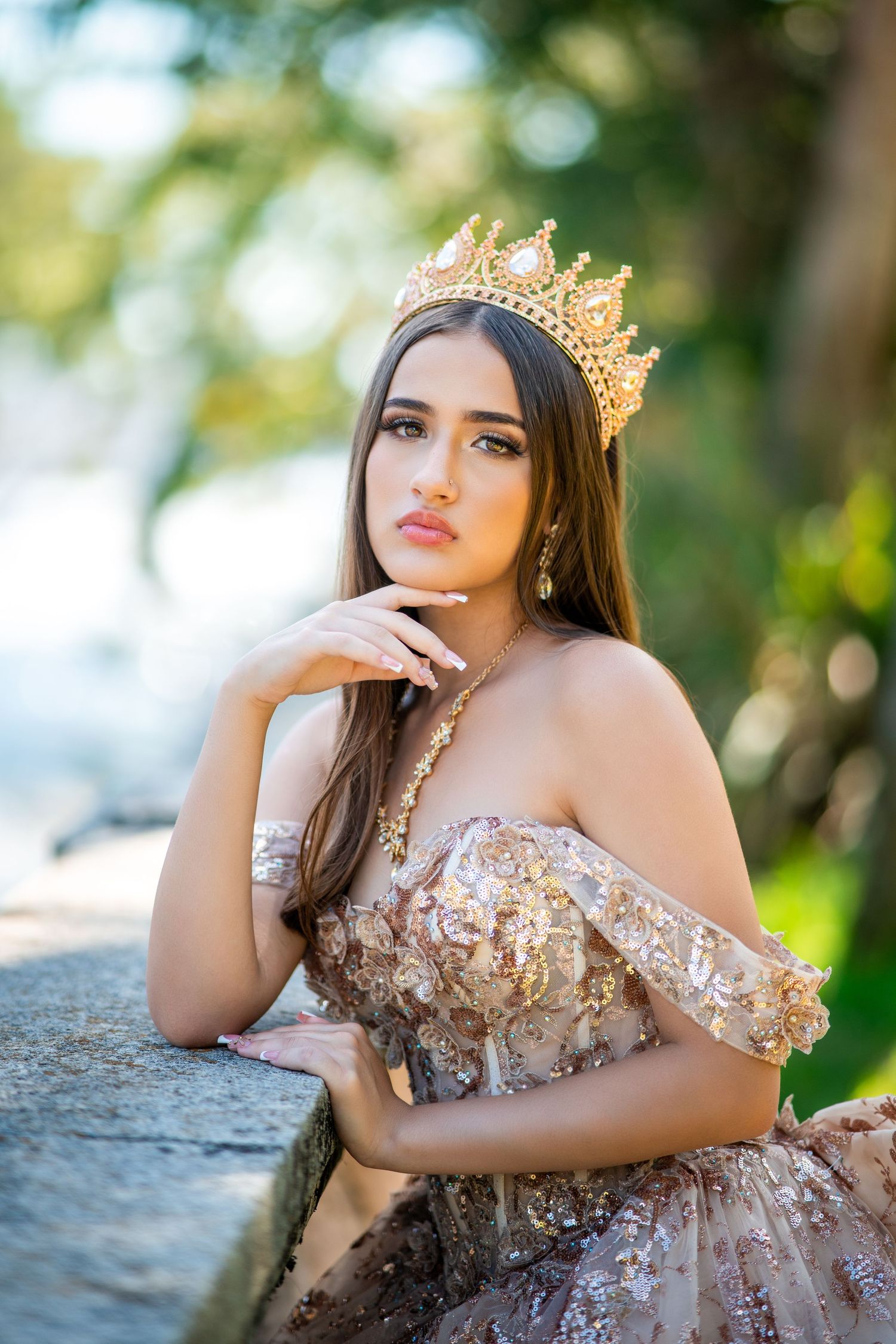 A young woman in a glittering gold gown and crown poses outdoors with soft bokeh lighting in the background.