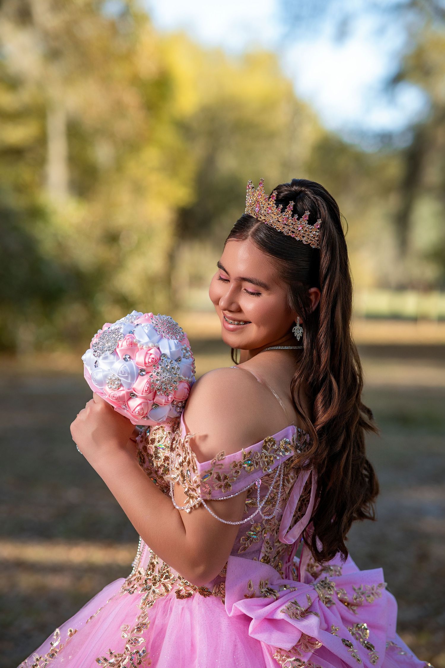 Young woman in pink quinceañera gown with gold crown holds floral bouquet outdoors, smiling gracefully.