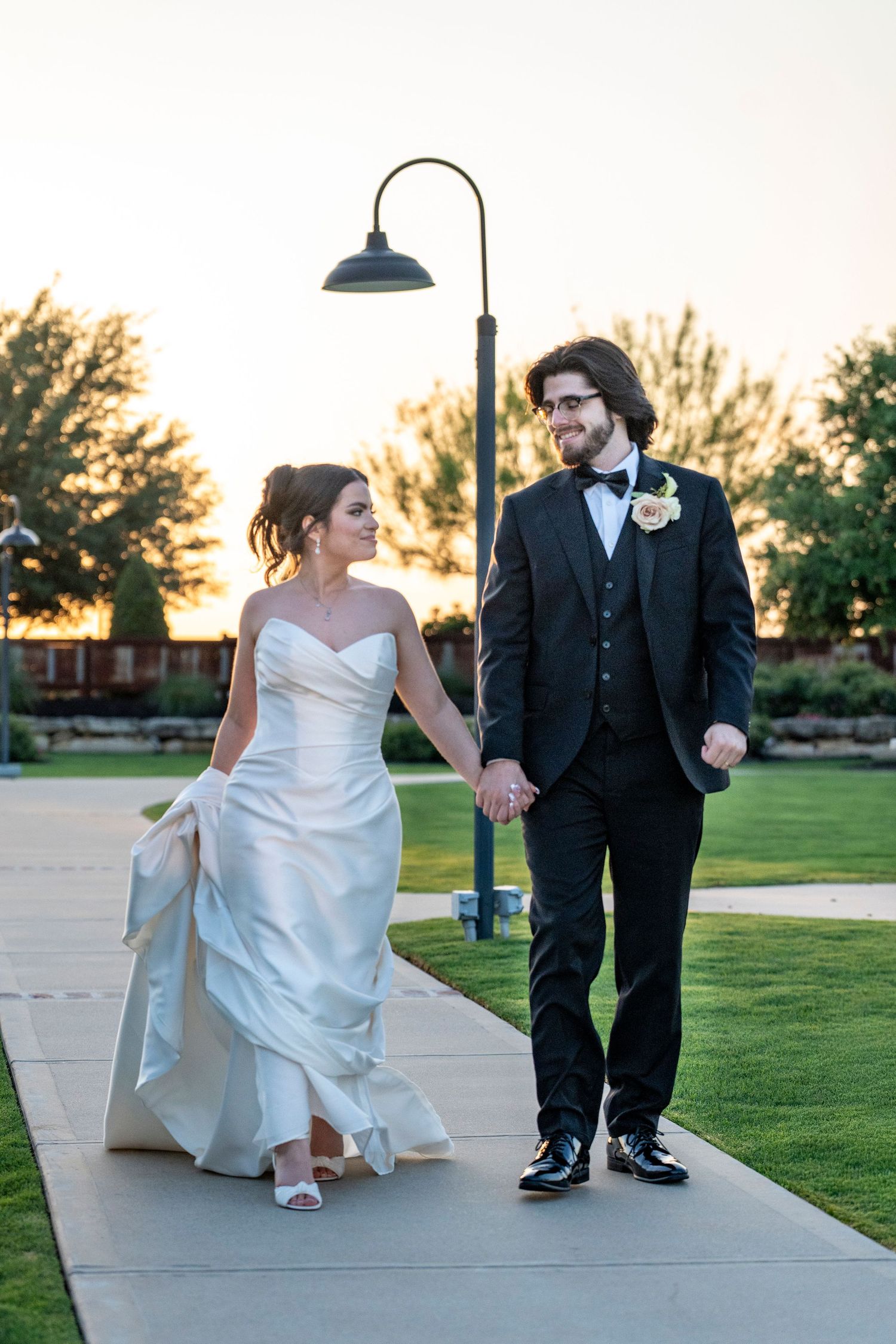 Bride and groom walk hand in hand at sunset along garden path in wedding attire.