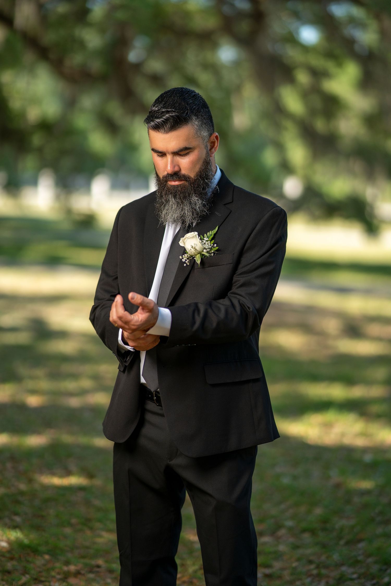 A bearded groom in a black suit with boutonniere adjusts his cufflinks outdoors under large oak trees.