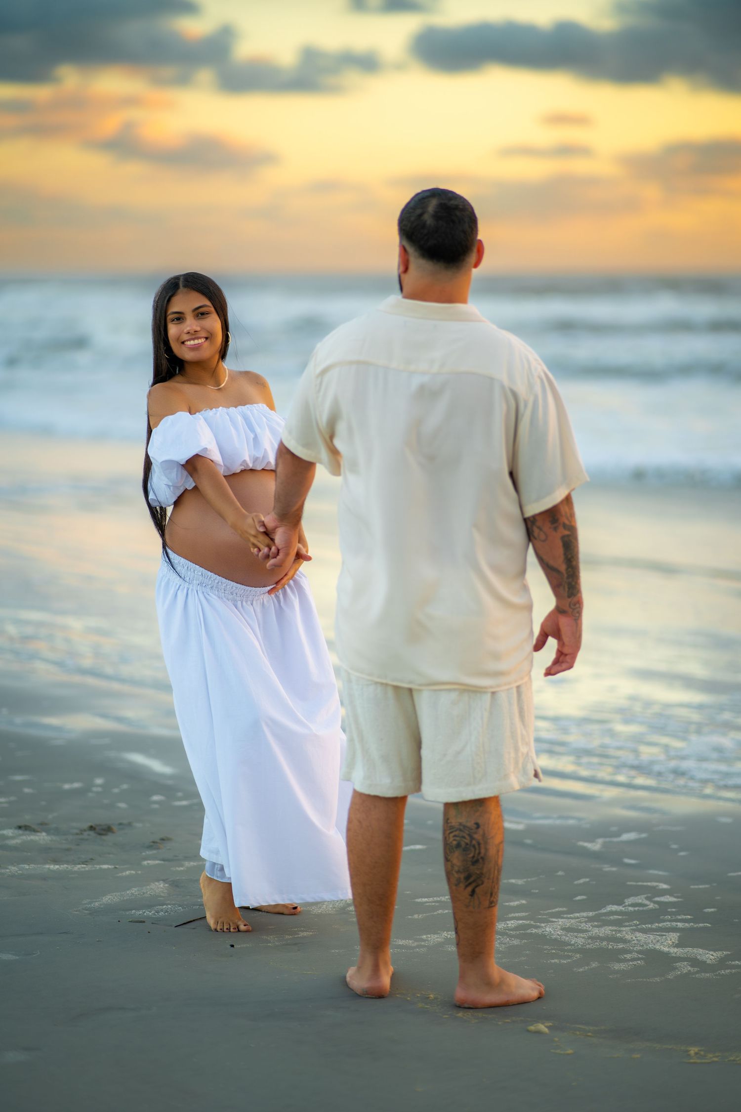 A couple in white clothing stands together on a beach at sunset while waves crash against the shore.