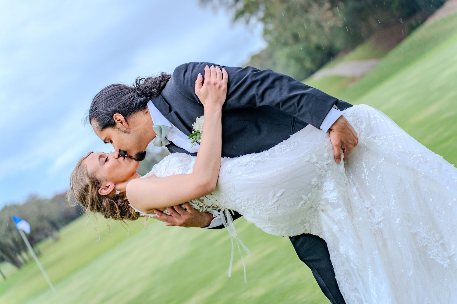 A couple in wedding attire shares a romantic kiss while dipping on a lush green lawn.