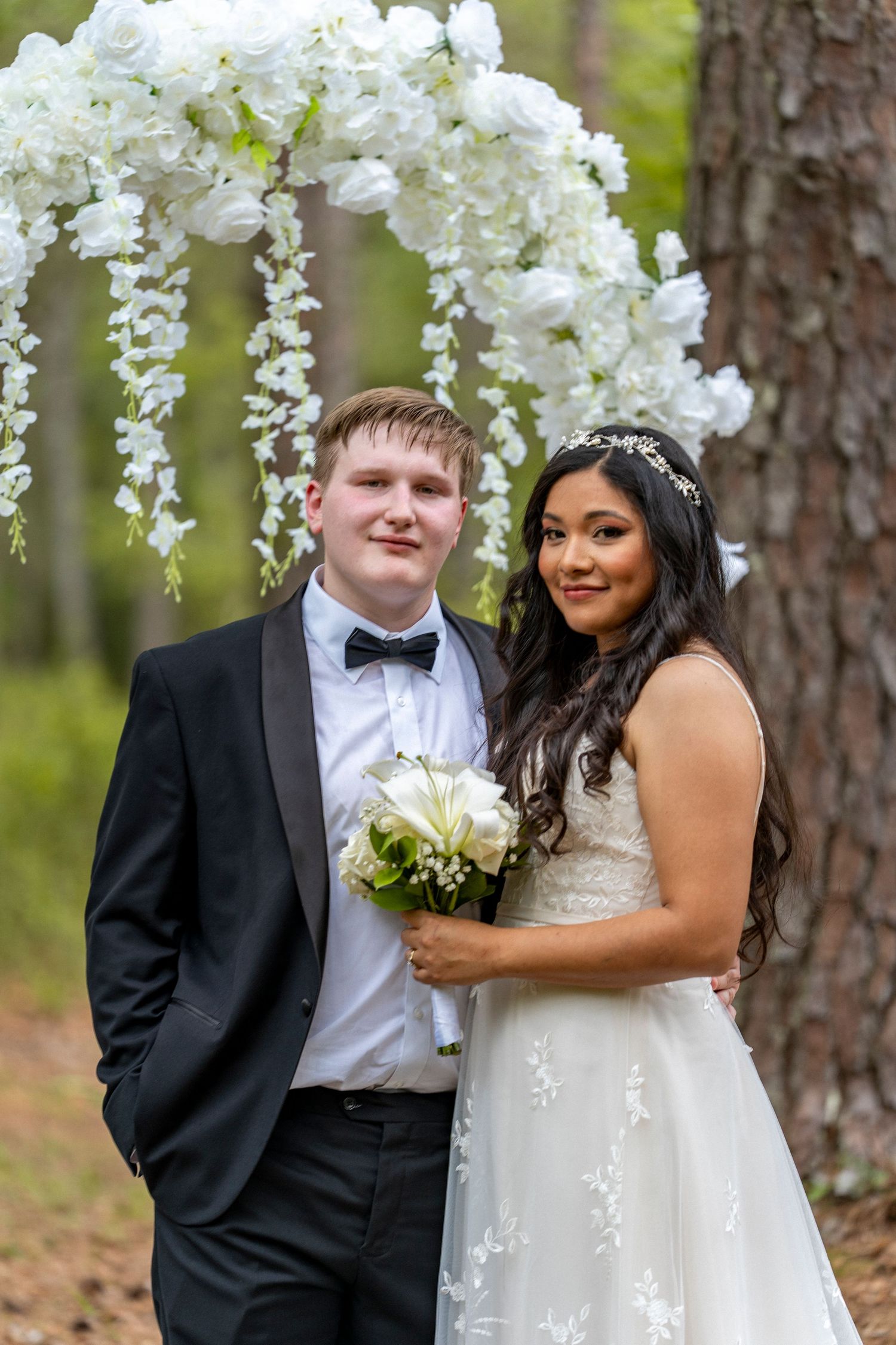 Bride and groom posing under white floral arch in forest, she holds bouquet in lace gown, he wears black tuxedo.