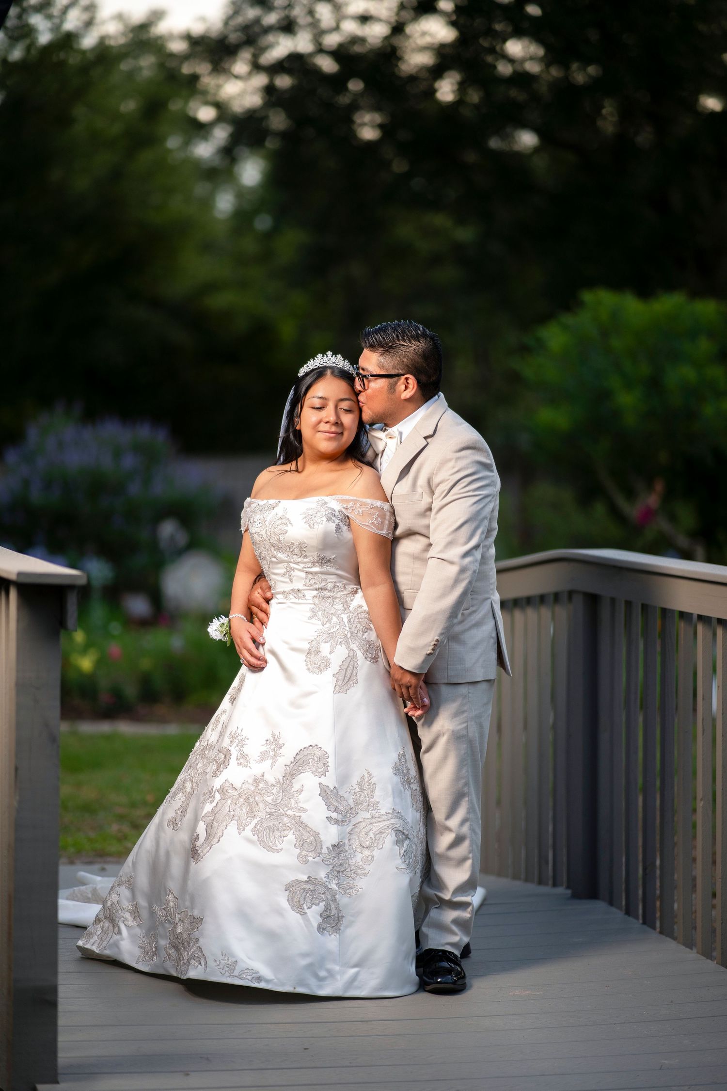Bride in white gown and groom in tan suit share a kiss on a garden bridge at golden hour.