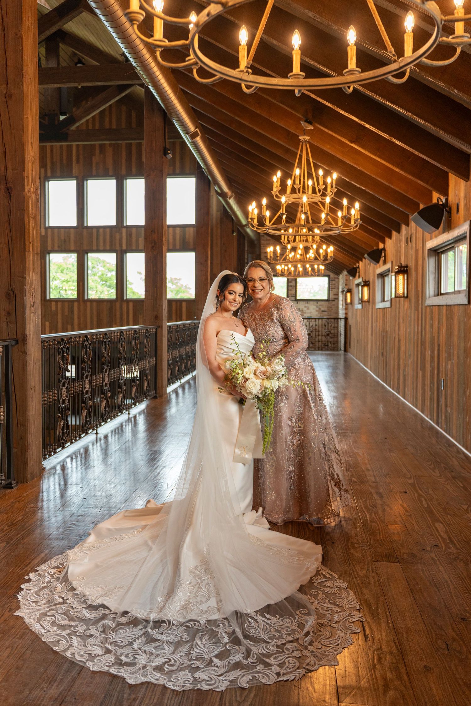 Bride in lace wedding gown with veil stands with mother in champagne dress under chandelier.