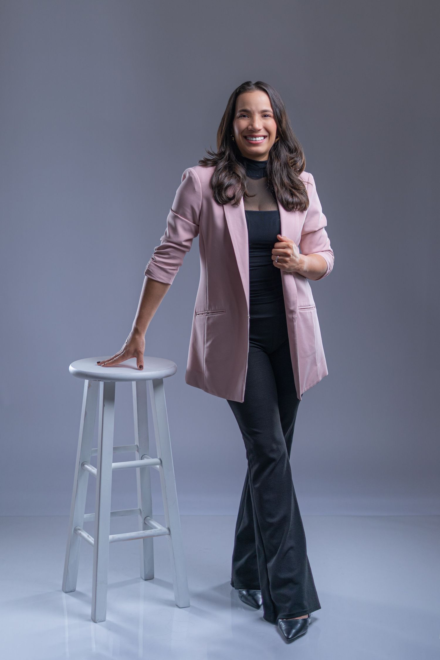 Professional wearing a pink blazer and black outfit poses confidently with a white stool against a gray backdrop.