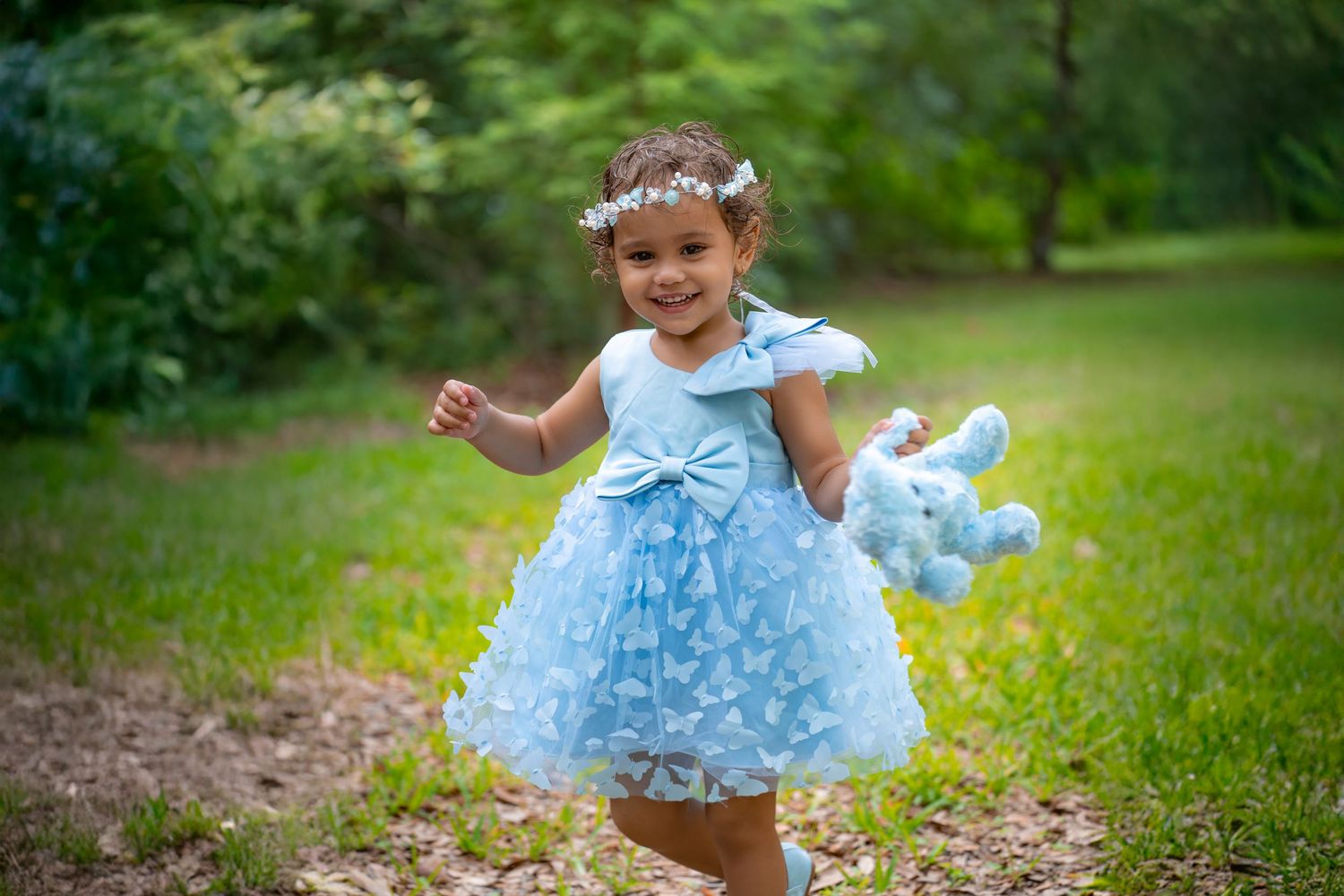 A child wearing a light blue dress with floral details twirls in a green outdoor garden setting.