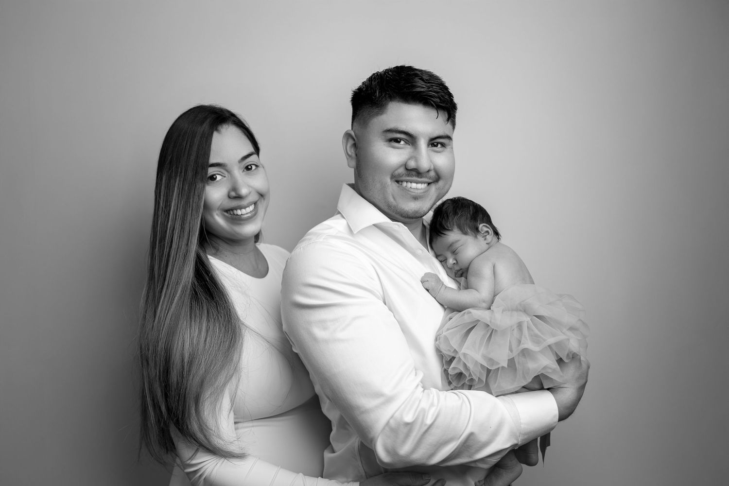 Black and white portrait of a family posing together with their newborn baby in a studio setting.