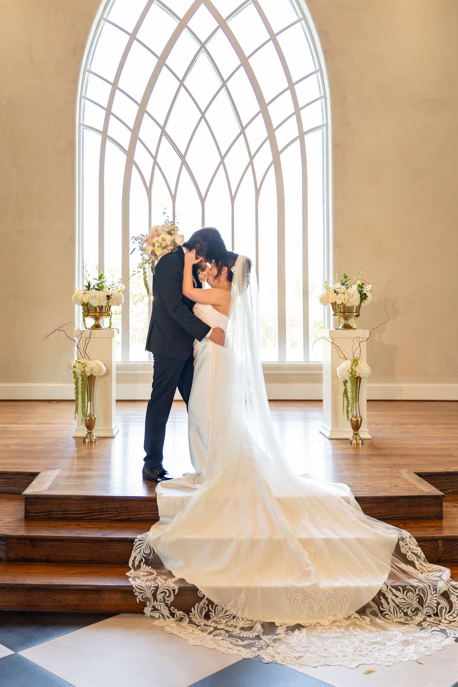 Bride and groom share romantic kiss at chapel altar with arched window and white floral arrangements.