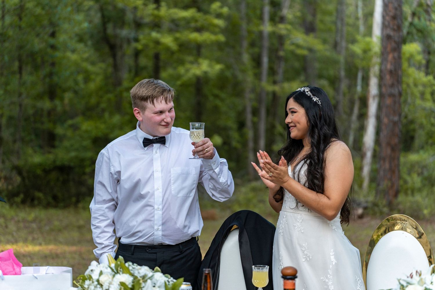 Bride and groom toasting with champagne glasses at outdoor wedding reception surrounded by lush green forest backdrop.