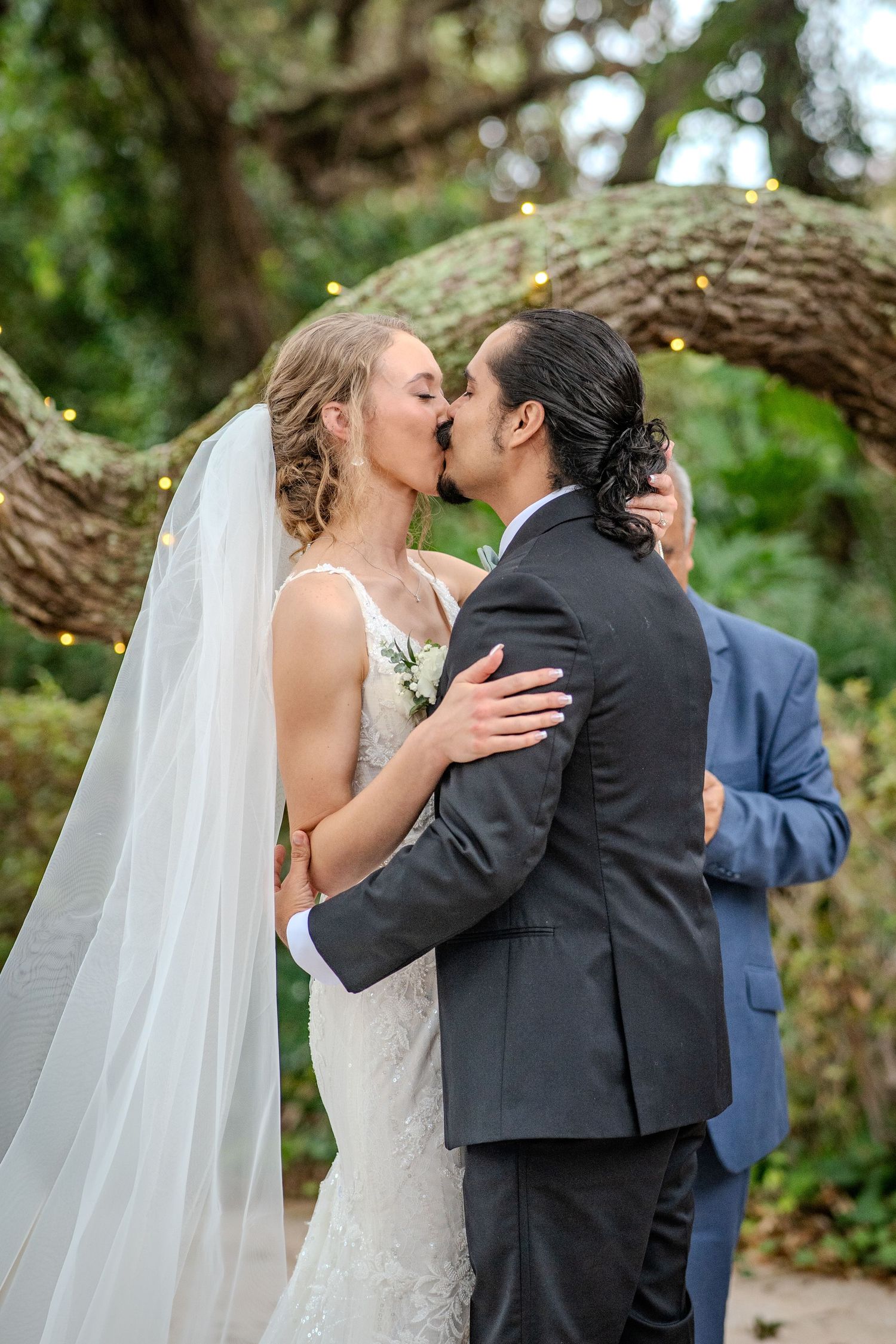A romantic wedding kiss sequence photographed outdoors under trees with a flowing white veil and formal attire.