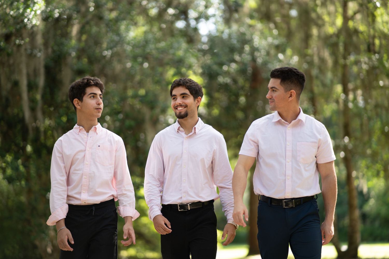 Groomsmen in light pink dress shirts stand together laughing and talking in an outdoor garden setting.