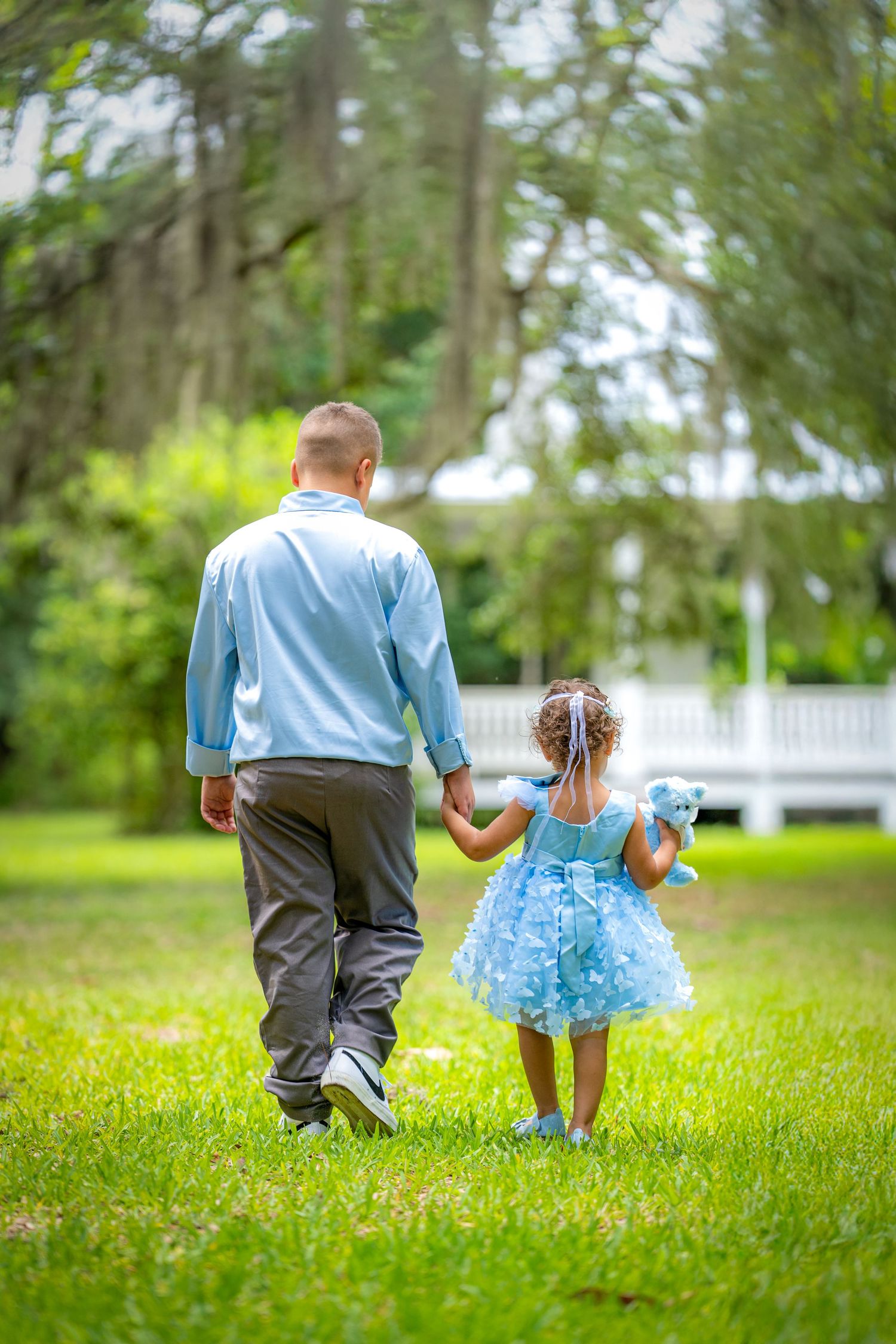 A person in light blue shirt walks hand-in-hand with small child wearing a puffy blue dress through a green garden setting.