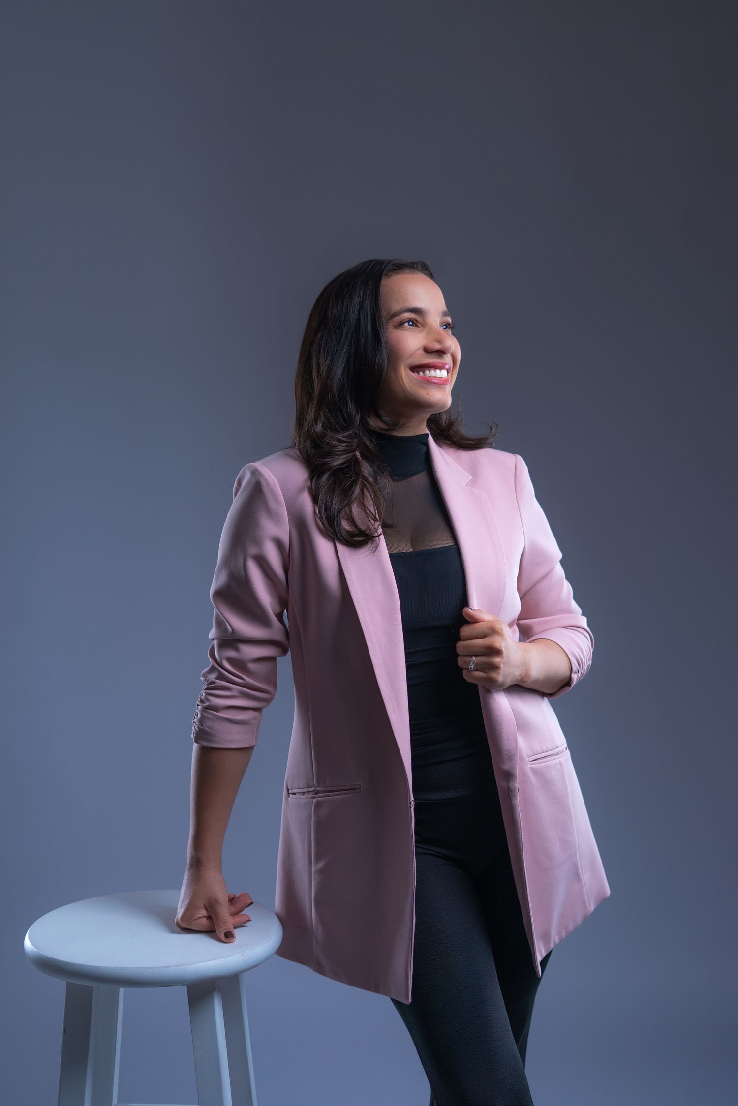 Professional portrait of someone wearing a pink blazer and black outfit leaning against a white stool against gray backdrop.