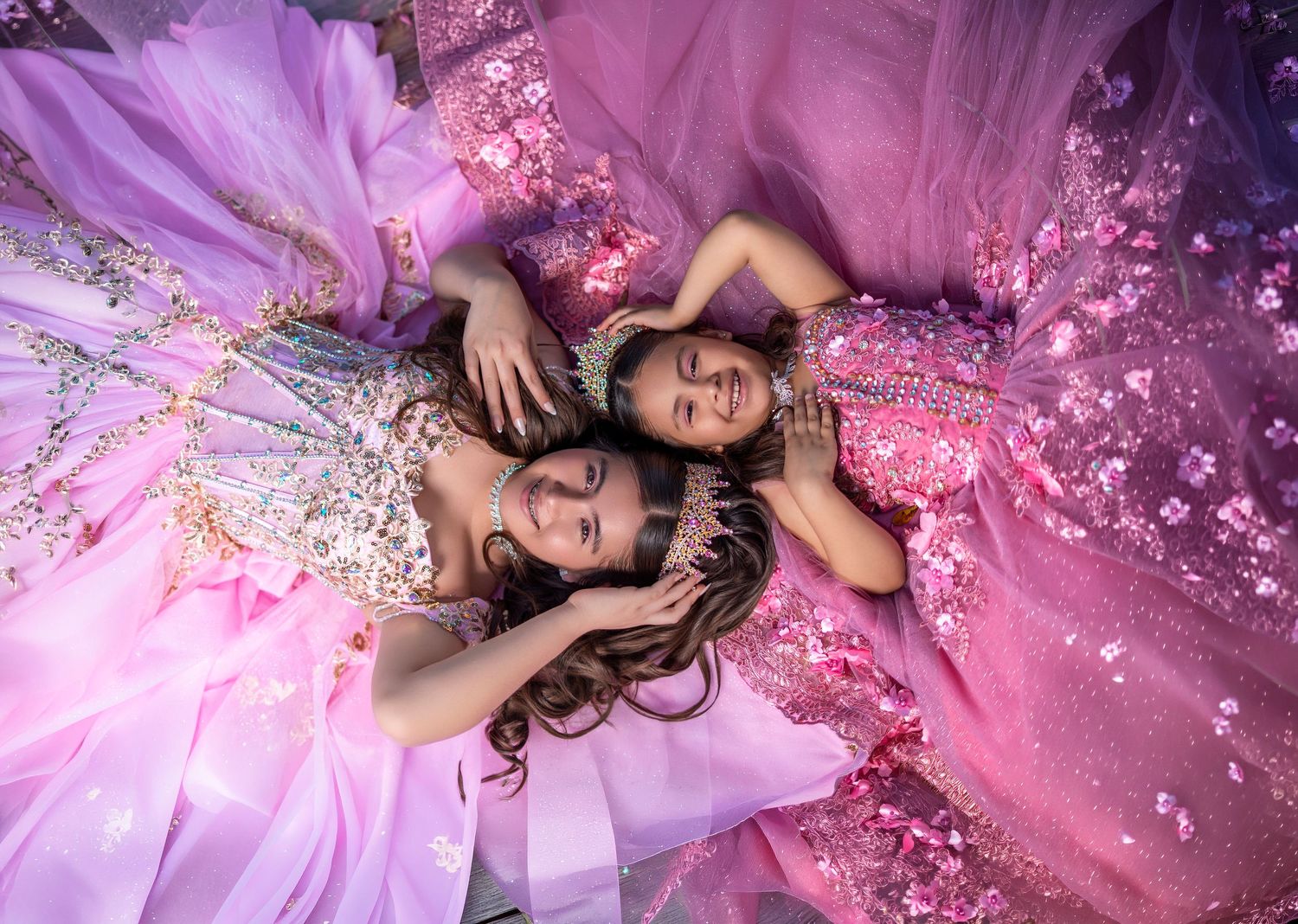 Two girls in pink and lavender princess gowns lying together surrounded by floral tulle fabric.