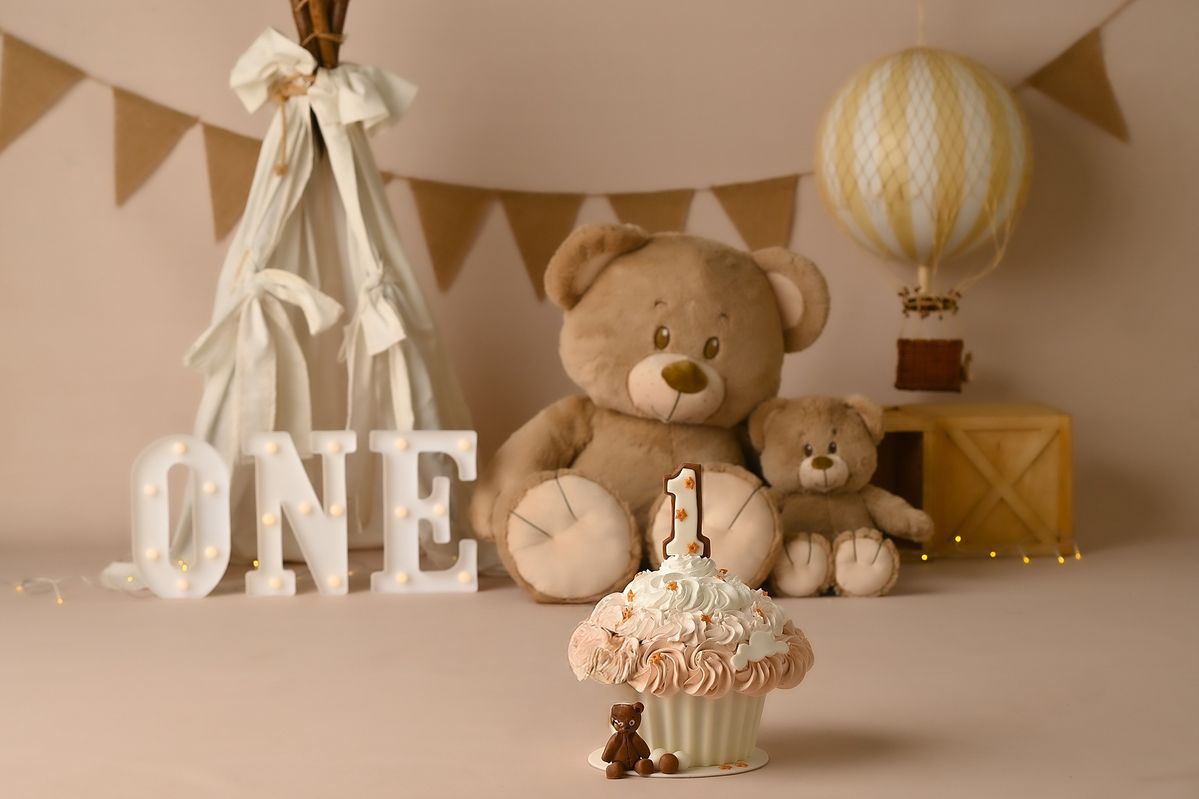First birthday party setup with teddy bears, hot air balloon decor, and white 'ONE' letters against neutral backdrop.