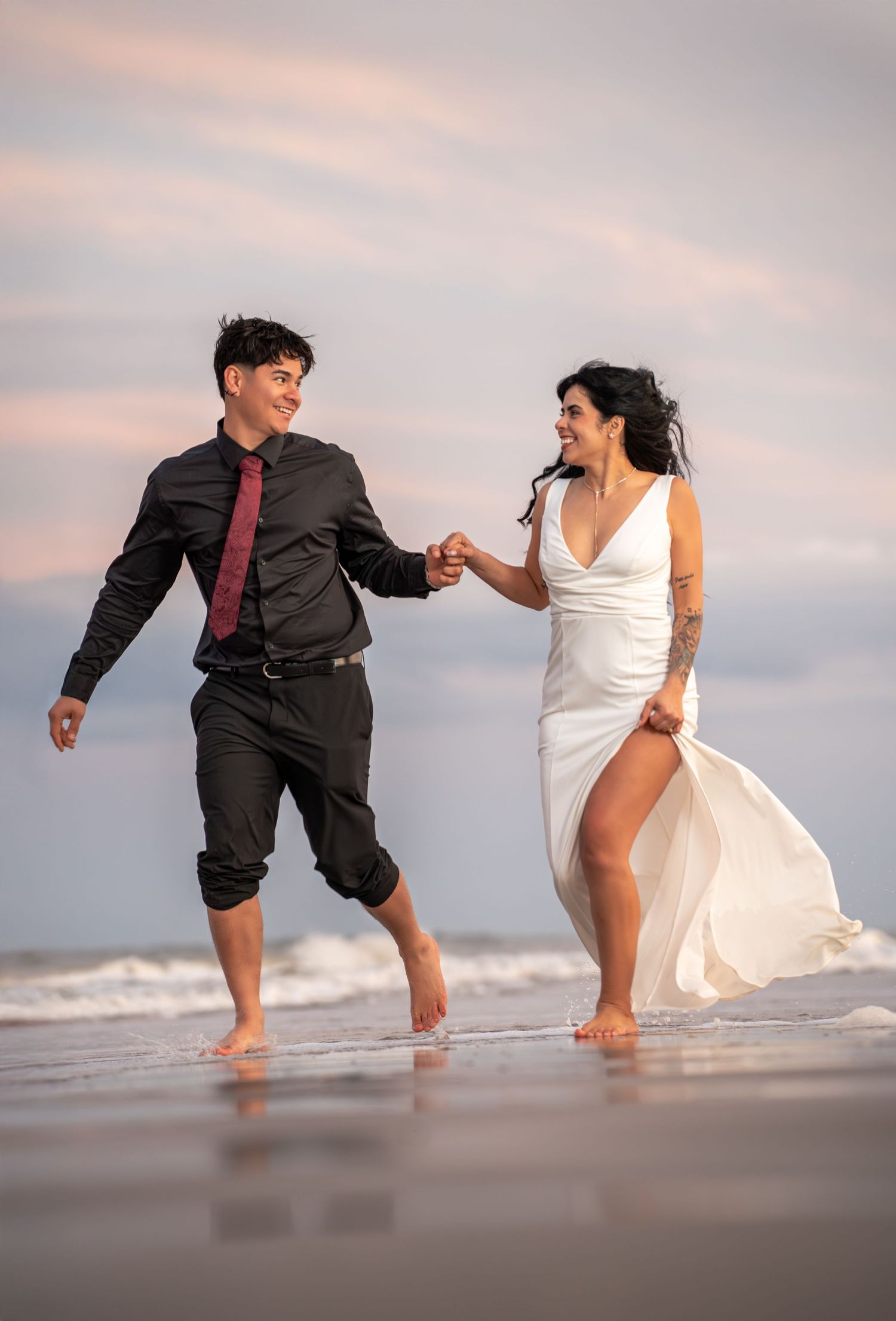 A couple playfully splashes and dances in the ocean waves at sunset wearing formal attire on the beach.
