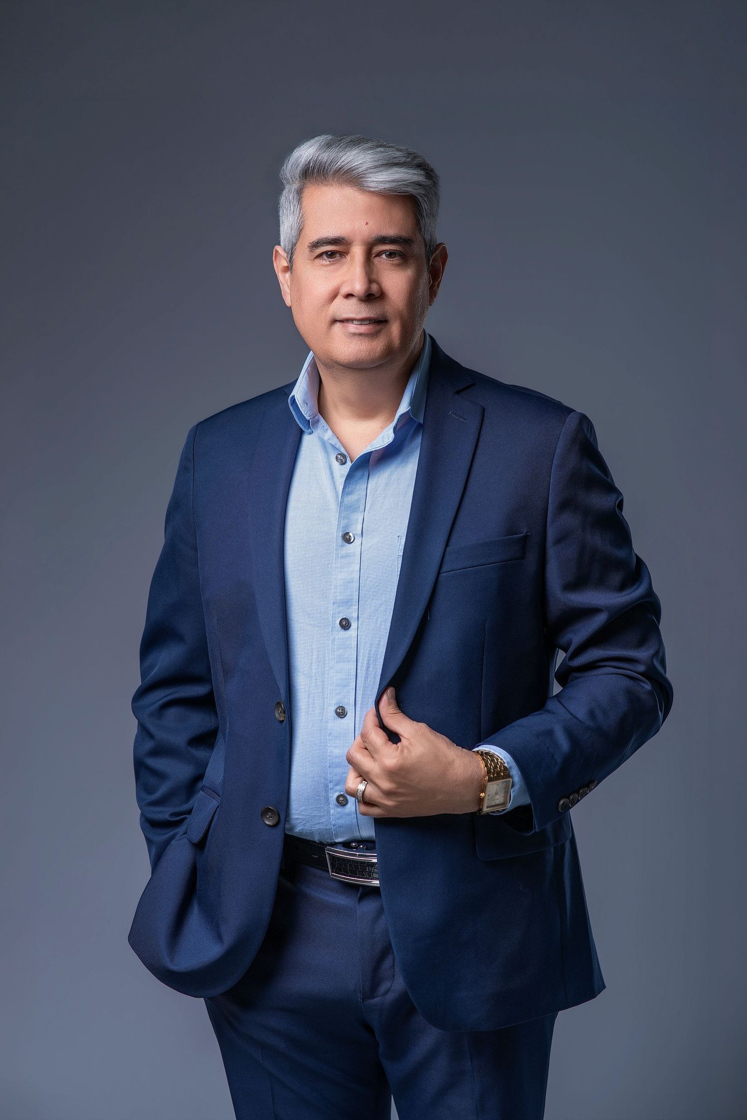 Distinguished middle-aged man with gray hair wearing navy blue suit and watch posing on gray background.