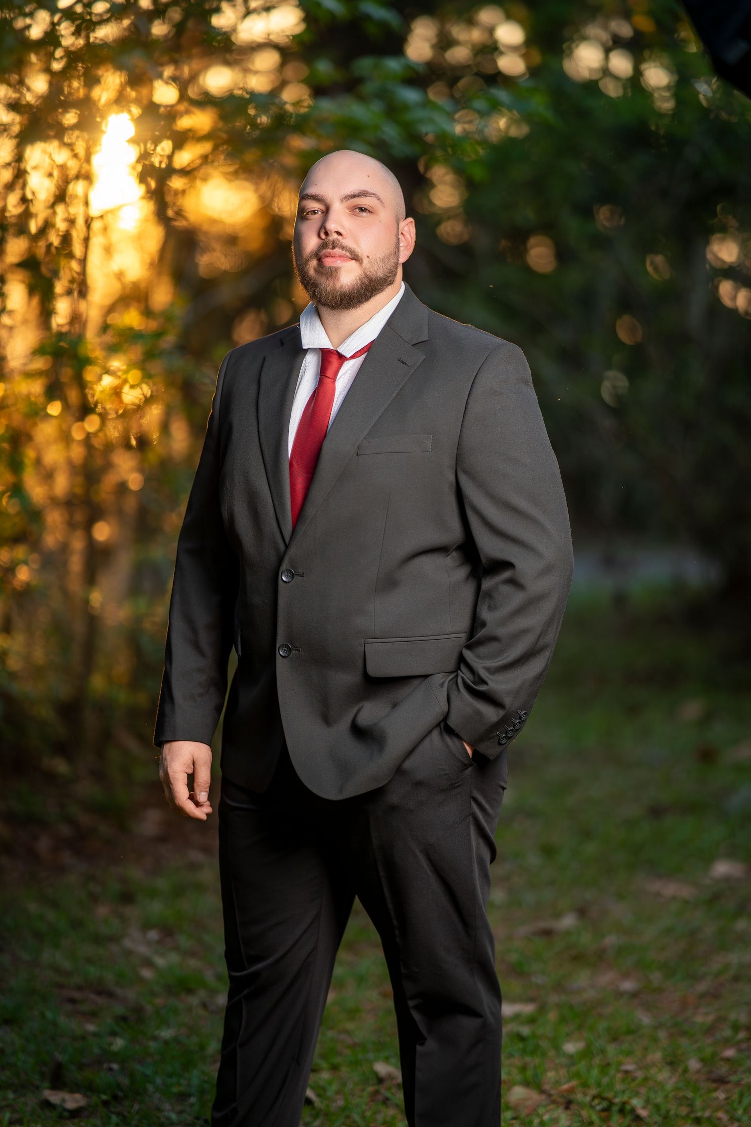 Bald man with beard in dark suit and red tie standing outdoors at golden hour sunset in forest setting.