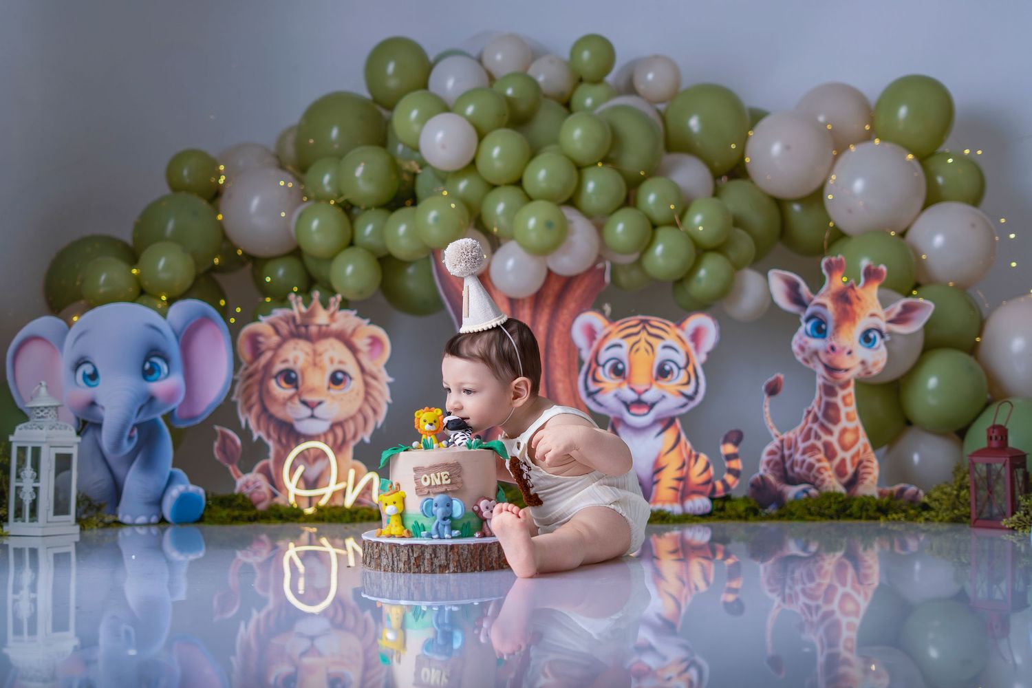 Baby's first birthday cake smash with jungle animal cutouts, green and white balloon arch, and colorful decorated cake.