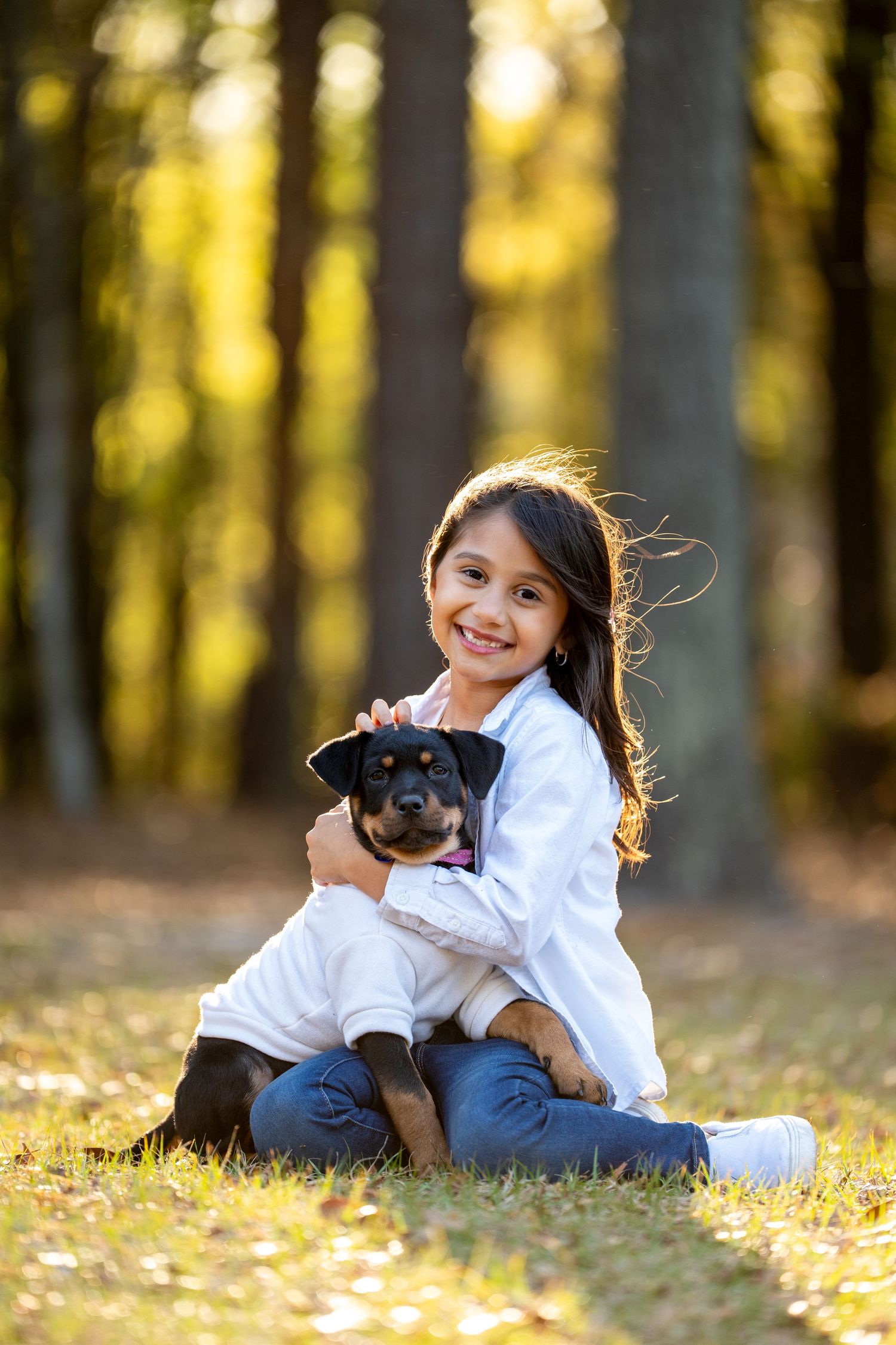 A young girl in jeans and white shirt plays with a black dog in autumn forest bathed in golden sunlight.