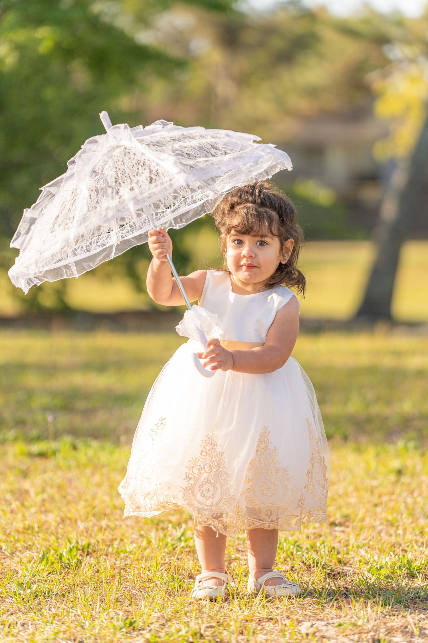 A small child in a white dress twirls a delicate lace parasol while playing in autumn leaves at sunset in a park.