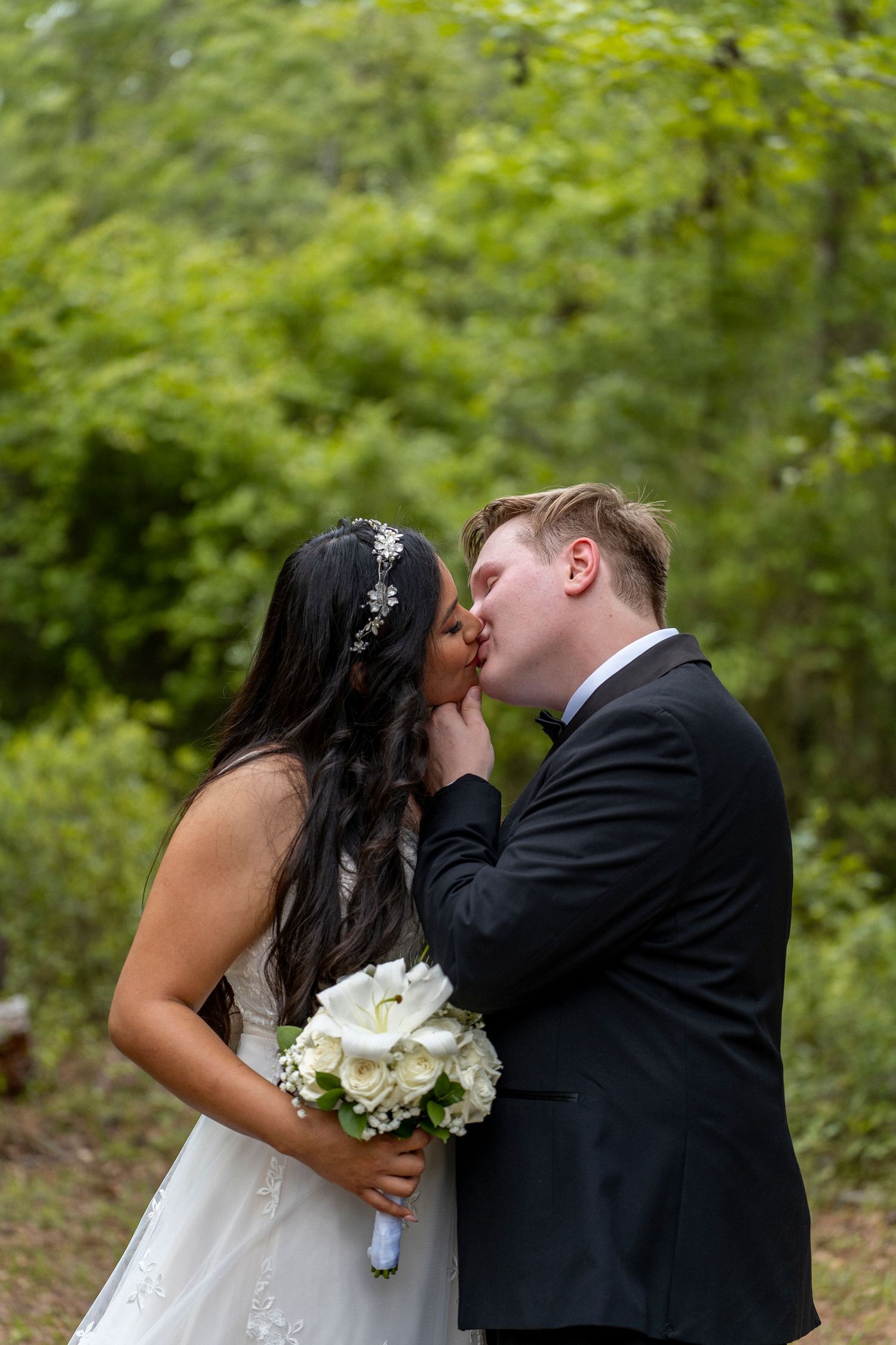 Bride and groom sharing first kiss outdoors, bride holds white rose bouquet wearing lace gown and floral headpiece.