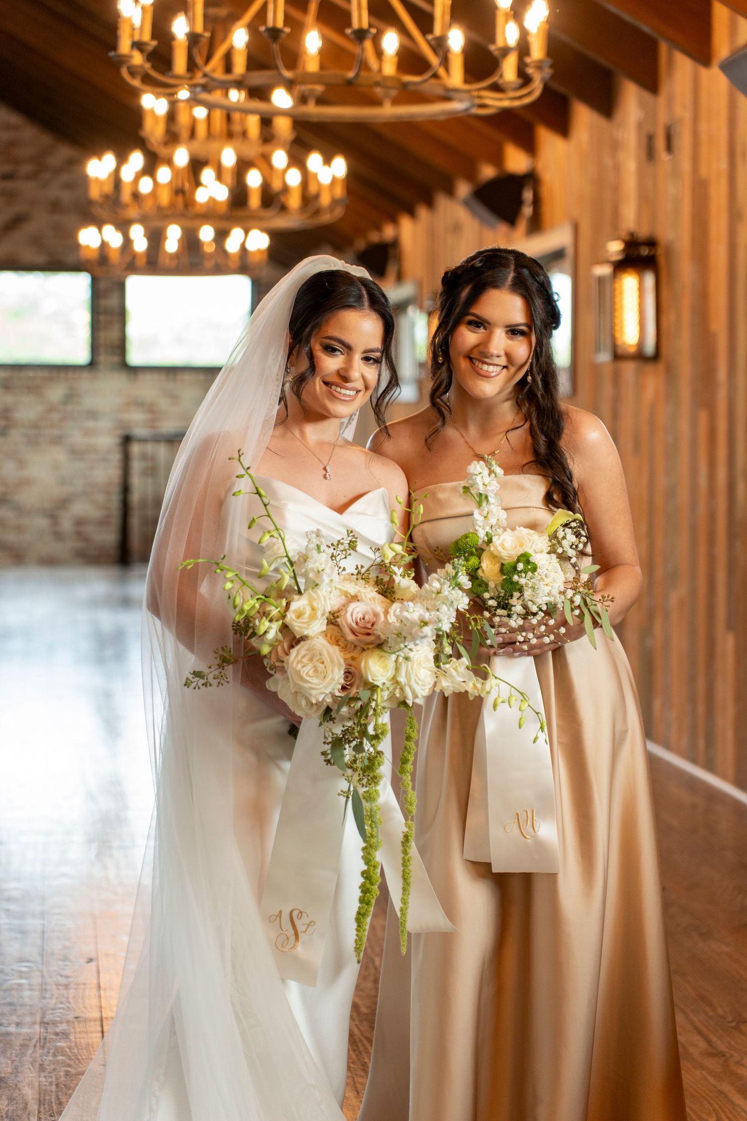 Bride in white gown and veil poses with bridesmaid in champagne dress, both holding white floral bouquets indoors.