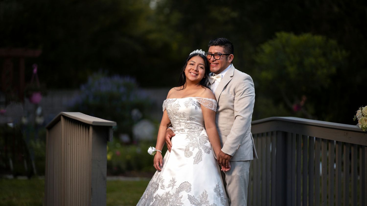 Elegant bride and groom pose together on a wooden bridge surrounded by lush garden greenery.