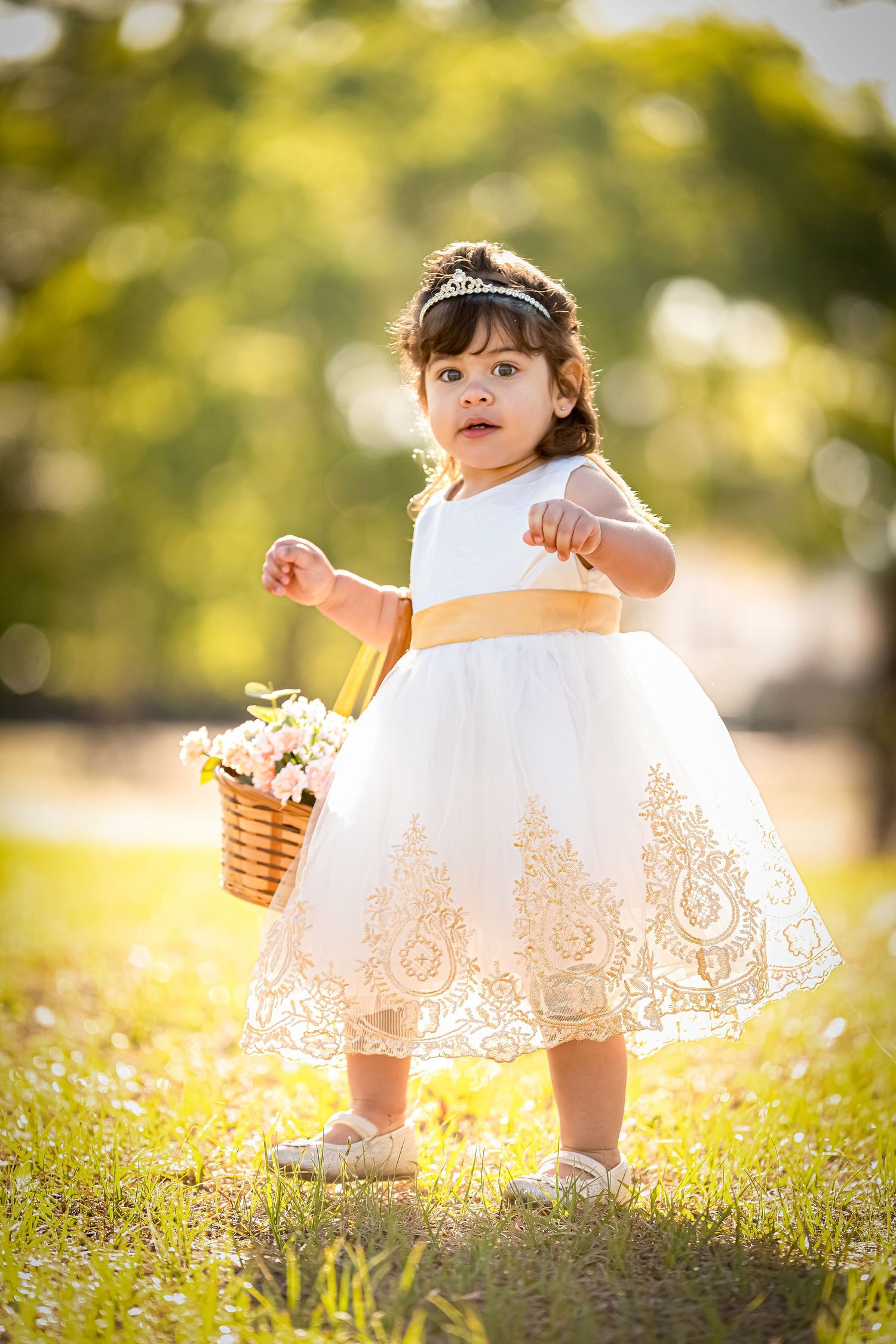 A small child in a white lace dress with orange sash holds a basket in a sunny garden setting.