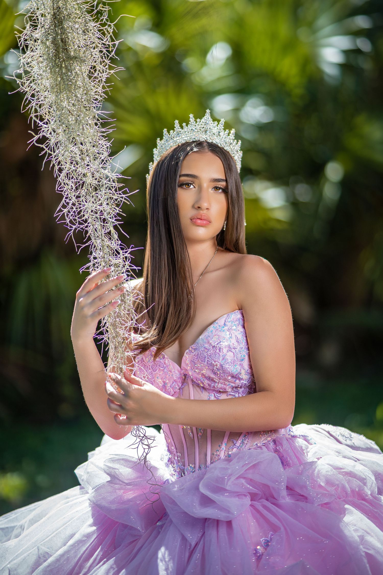 A person in a lavender princess gown with crown poses outdoors holding Spanish moss against tropical foliage.